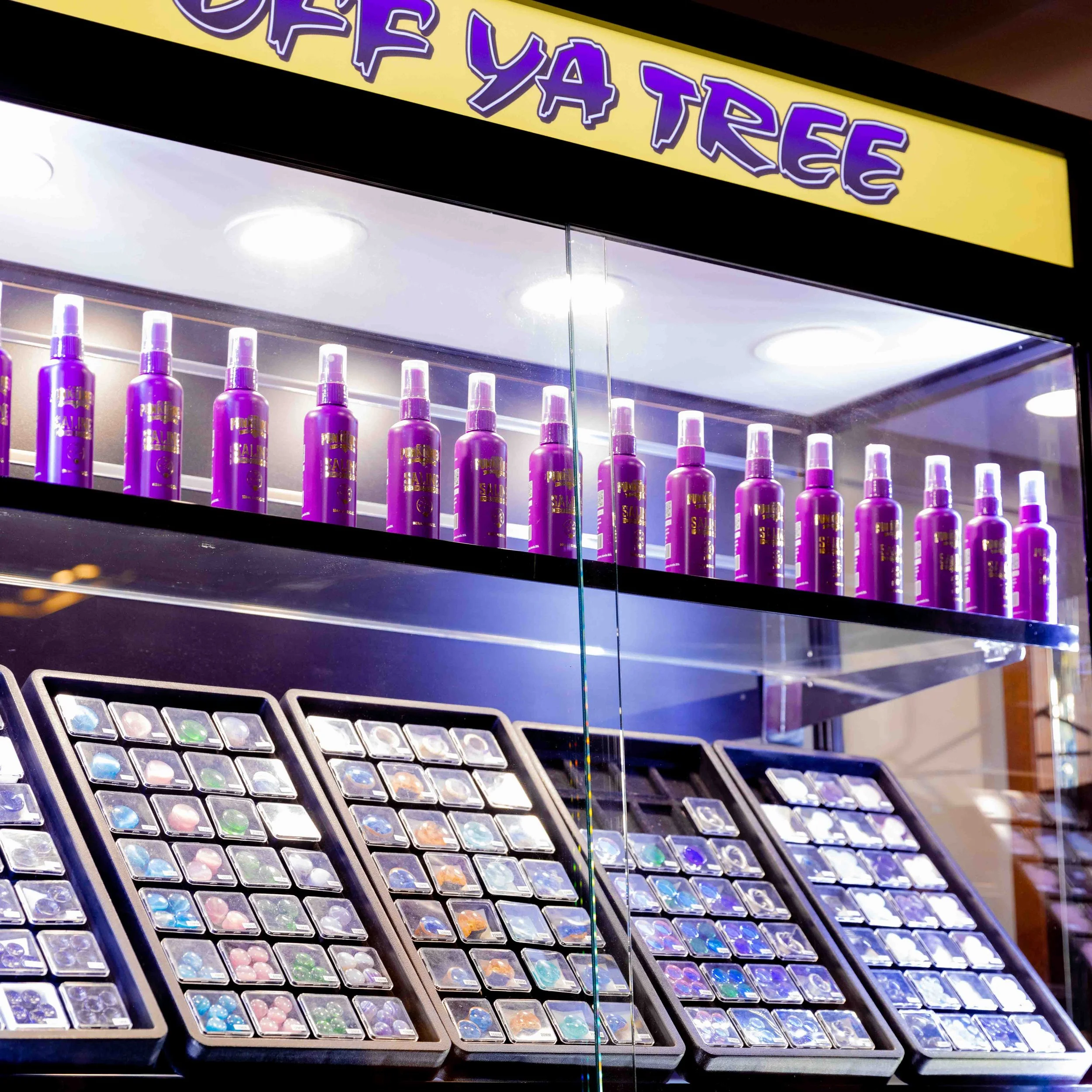 Display of purple spray bottles labeled "Purple Ya Tree" on a glass shelf, with colorful glitter nail polish sets in open cases below.