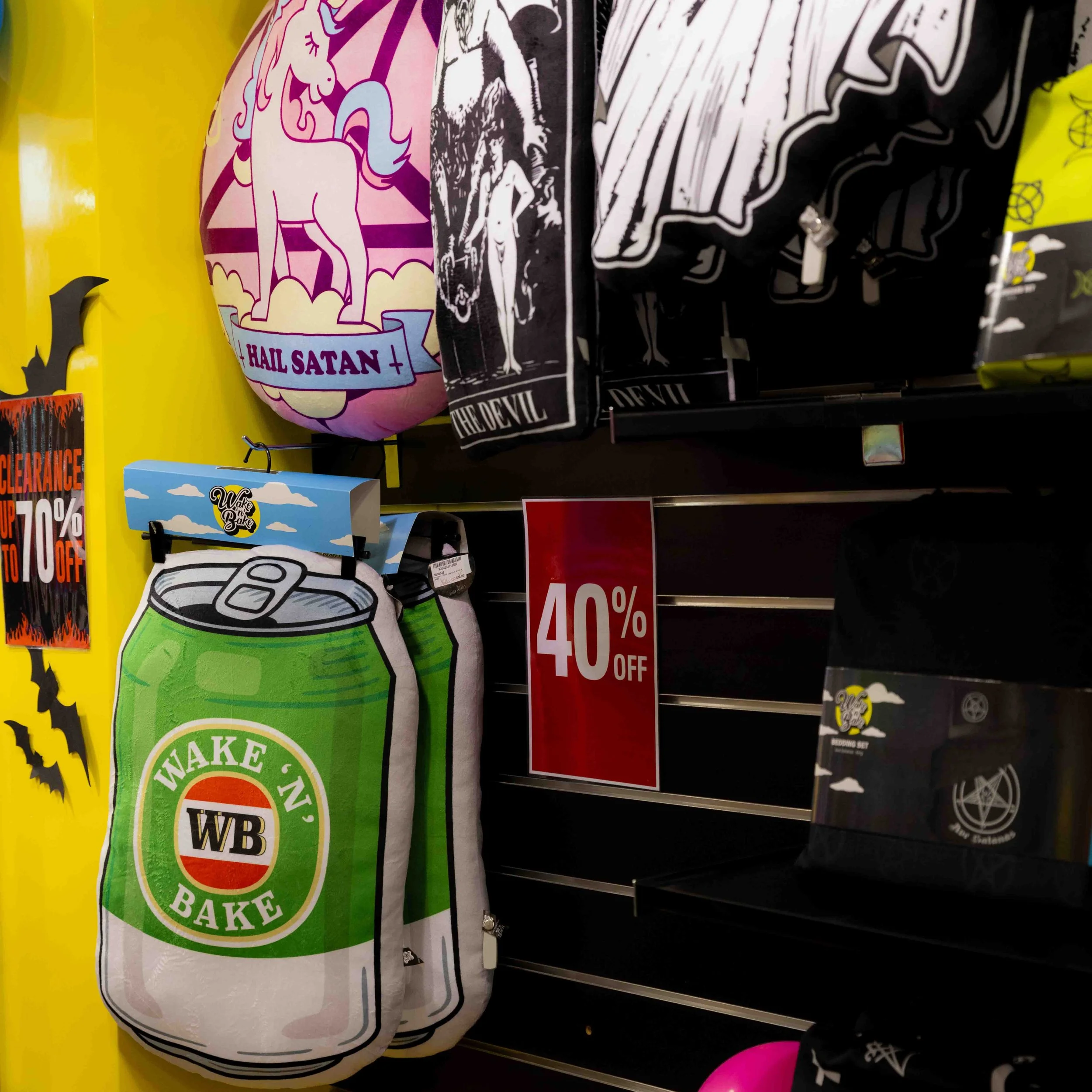 Halloween-themed merchandise on display, including black and white flags with spooky images, a balloon with a unicorn and the text 'Hail Satan,' and a plush beer can with the label 'Wake 'n Bake,' with a red sign offering 40% off.