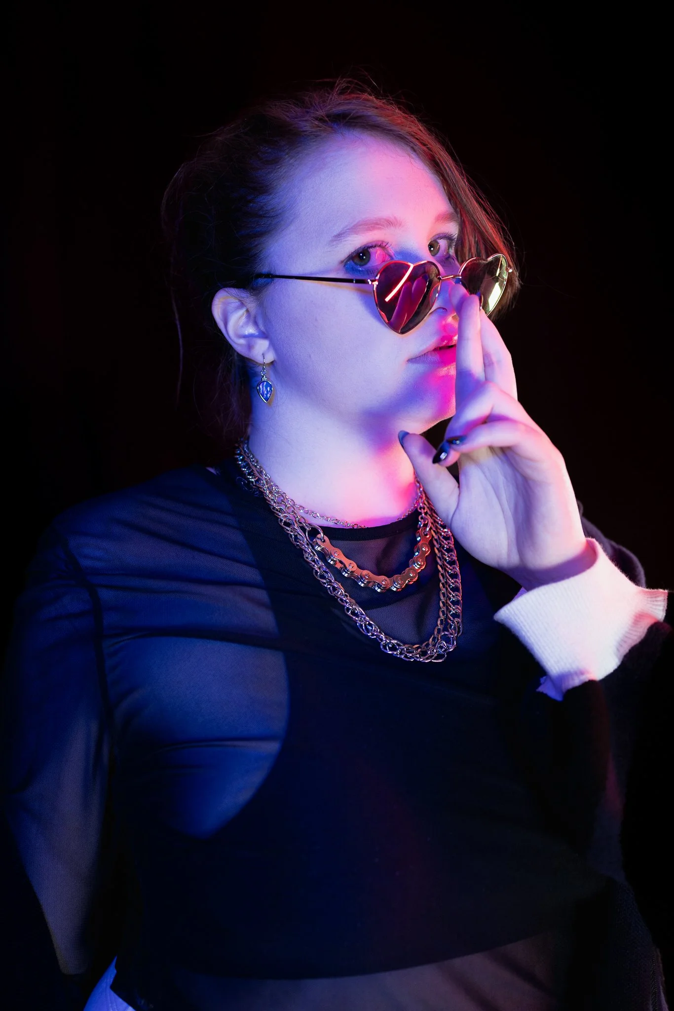 Young woman with pink and blue neon lighting, wearing heart-shaped sunglasses, layered gold necklaces, earrings, and a black top with sheer sleeves, holding her finger to her lips.