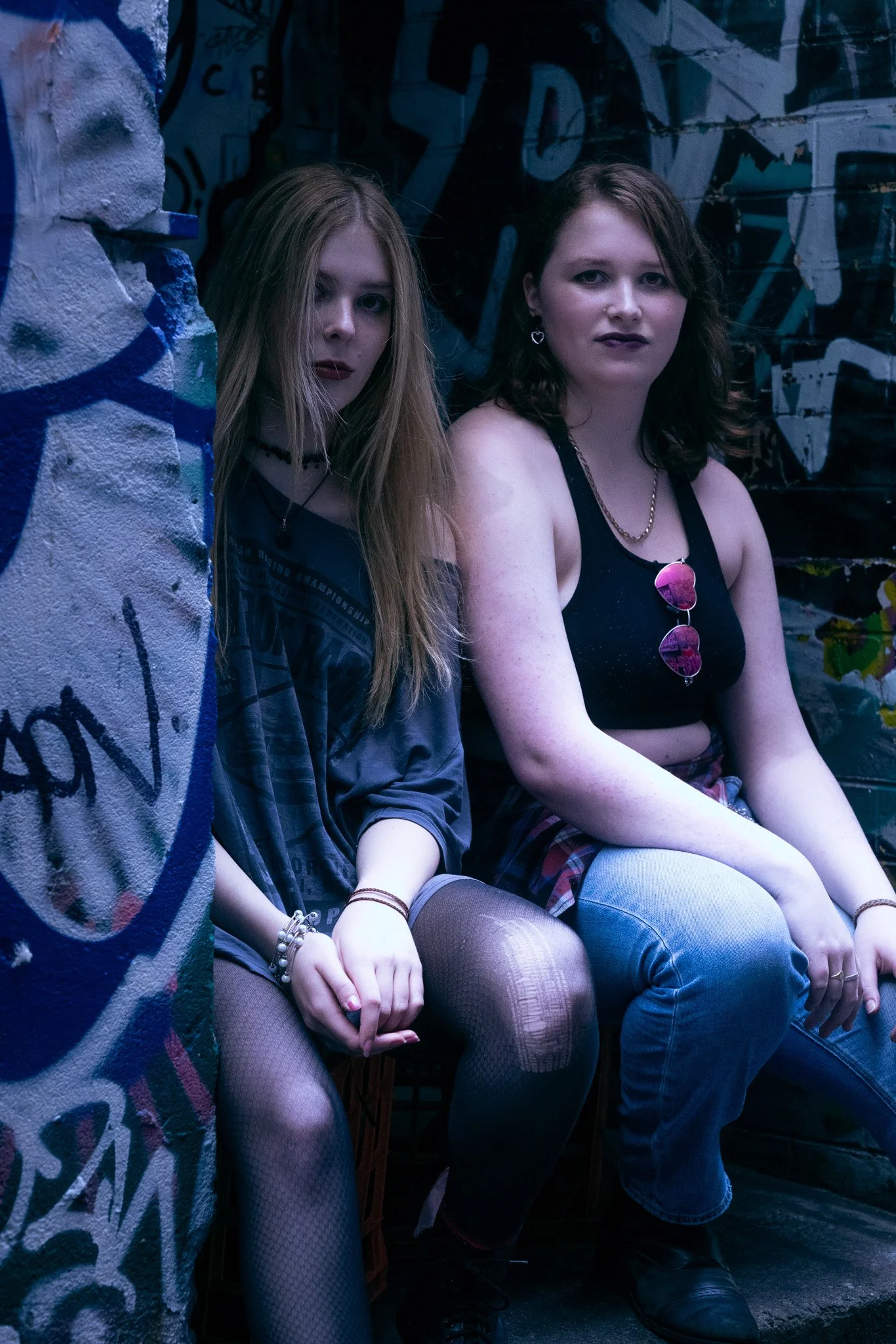 Two young women sitting in a graffiti-covered alleyway, looking at the camera. One has long blonde hair and wears fishnet tights, a choker, and silver bracelets; the other has shoulder-length dark hair, glasses hanging from a chain around her neck, a