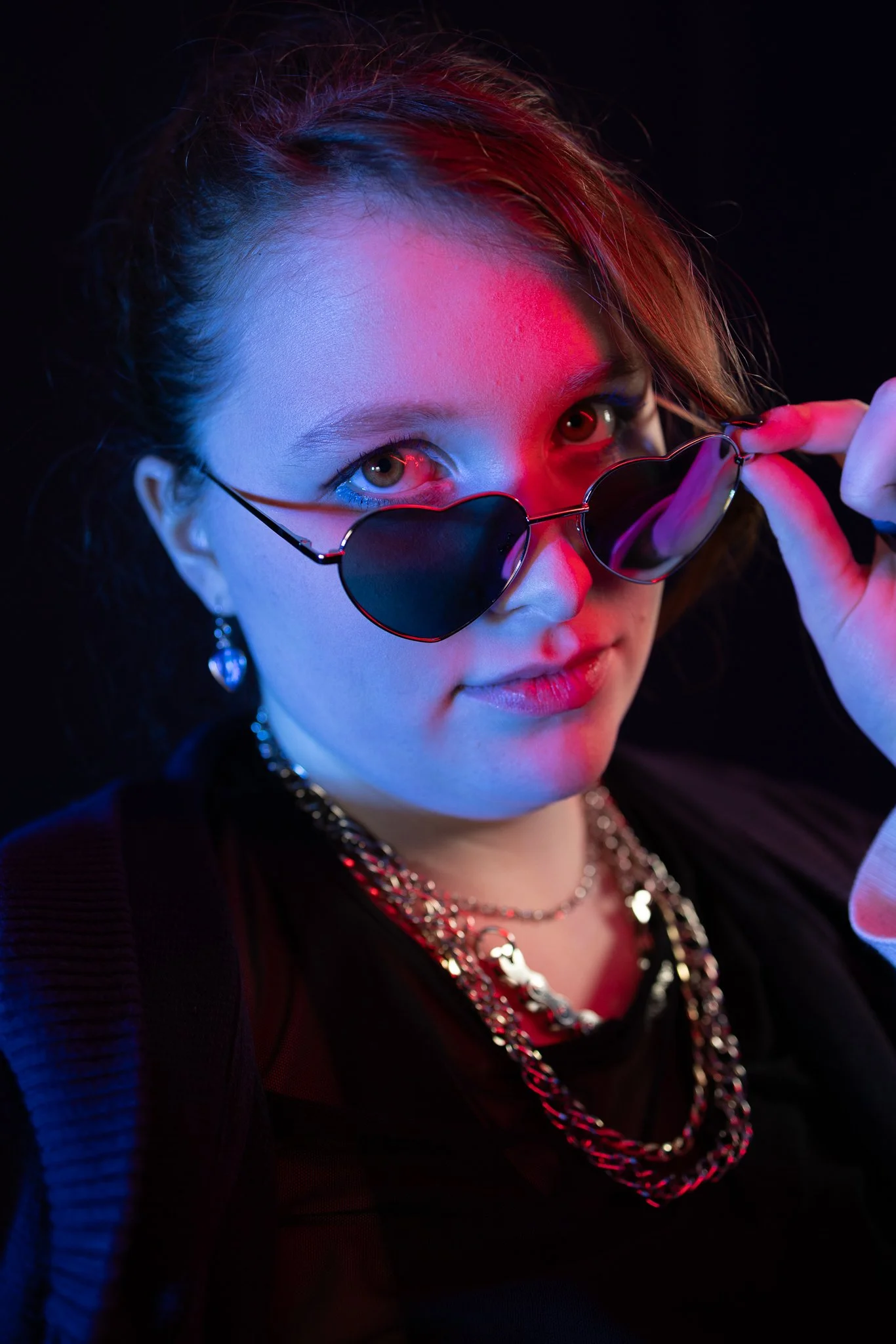 A young woman with light skin and brown hair, wearing heart-shaped sunglasses, layered necklaces, earrings, and dark clothing, posing against a dark background with pink and blue lighting.