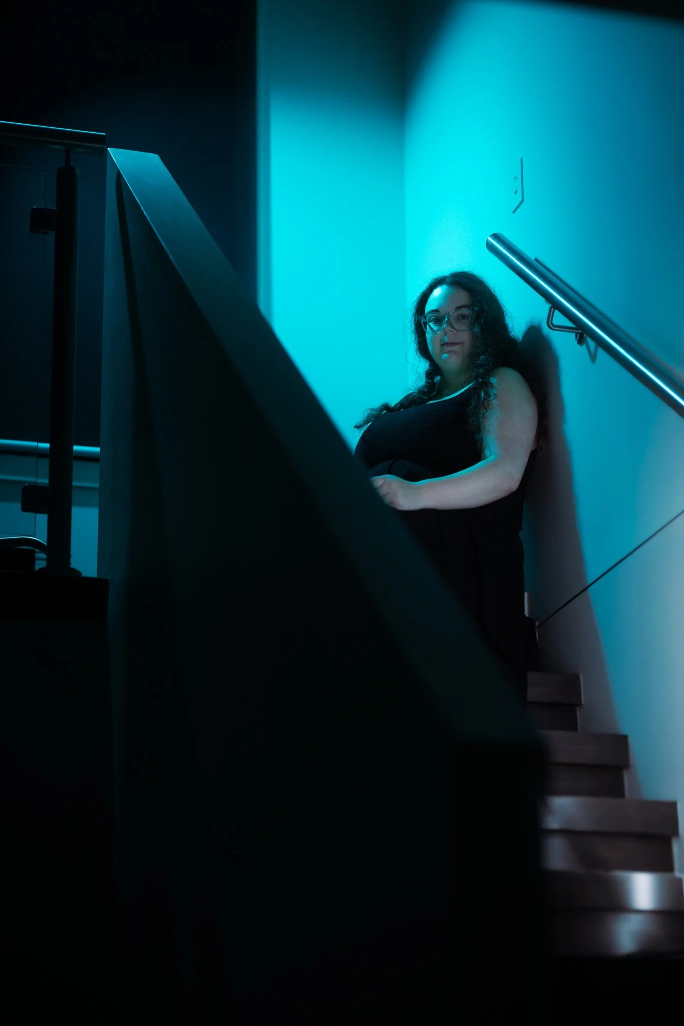 Young woman with long curly hair and glasses sitting on staircase in low lighting, illuminated by teal-blue light.