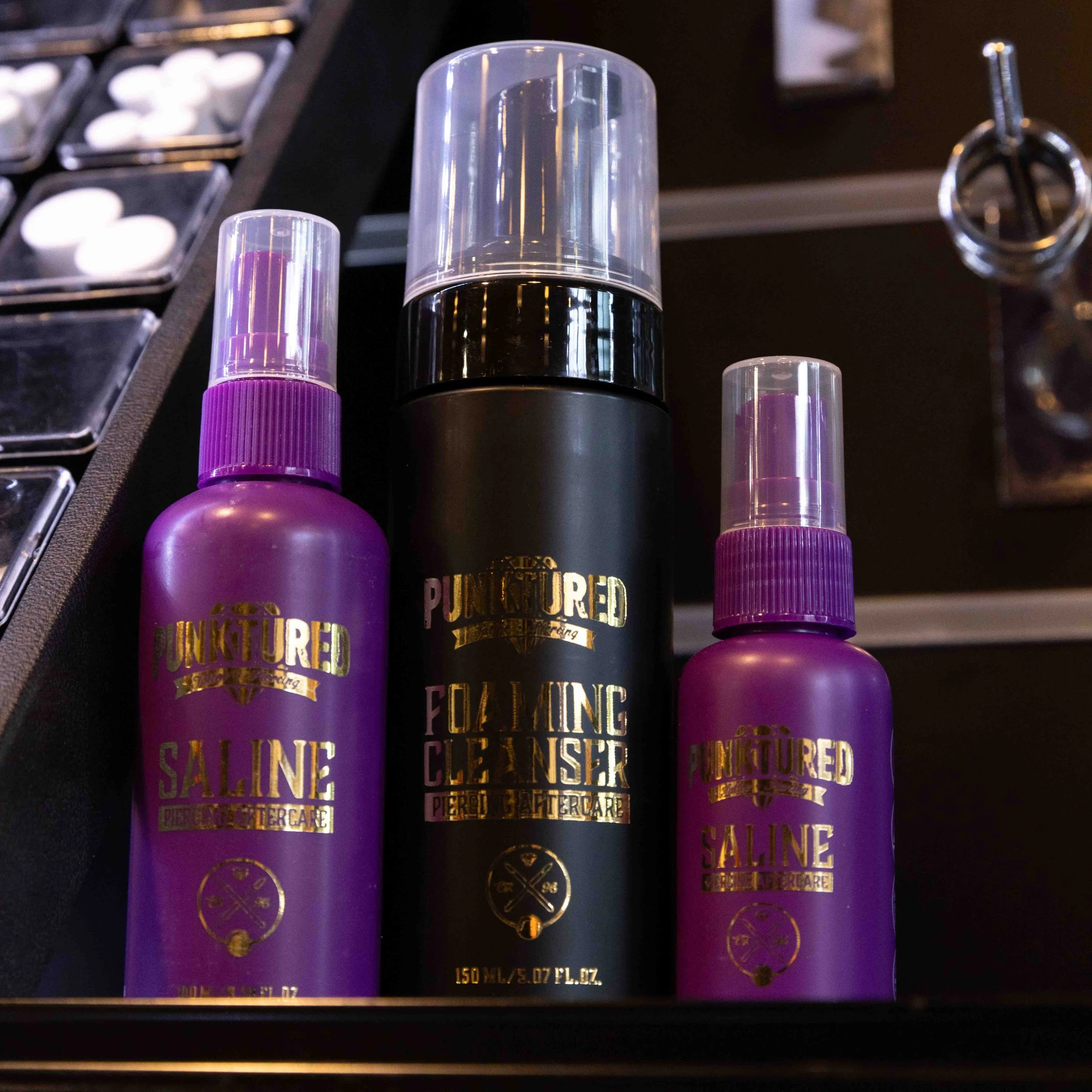 Three bottles of Punktured skincare products: two purple bottles labeled Saline, one black bottle labeled Foaming Cleanser, arranged on a dark surface.