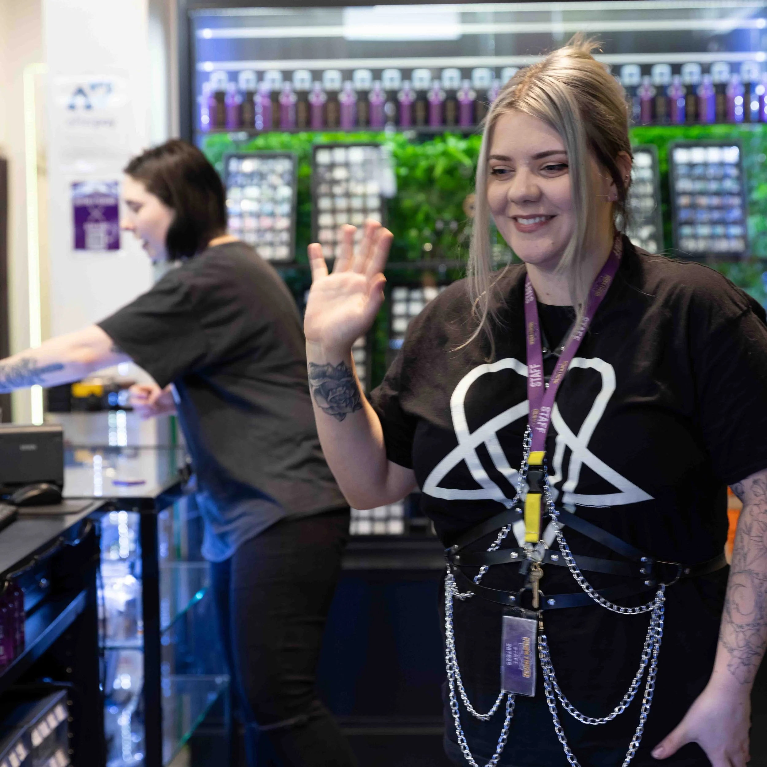 Two women working behind a counter, one smiling and waving at the camera, in a store with vape products in the background.