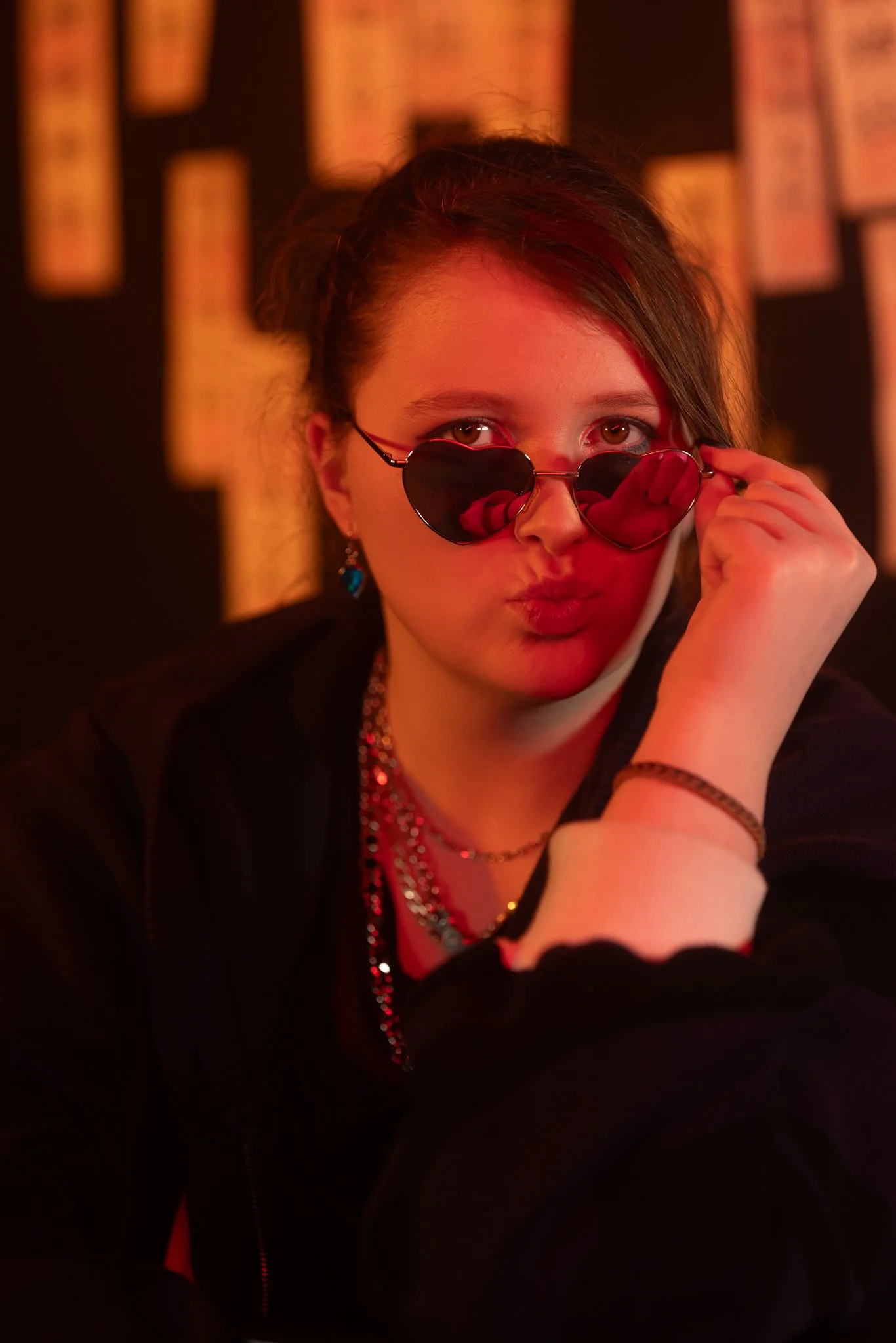 A young woman with dark hair and light skin wearing heart-shaped sunglasses, jewelry, and a black jacket, looking at the camera with a serious expression against a blurred background with warm lighting.