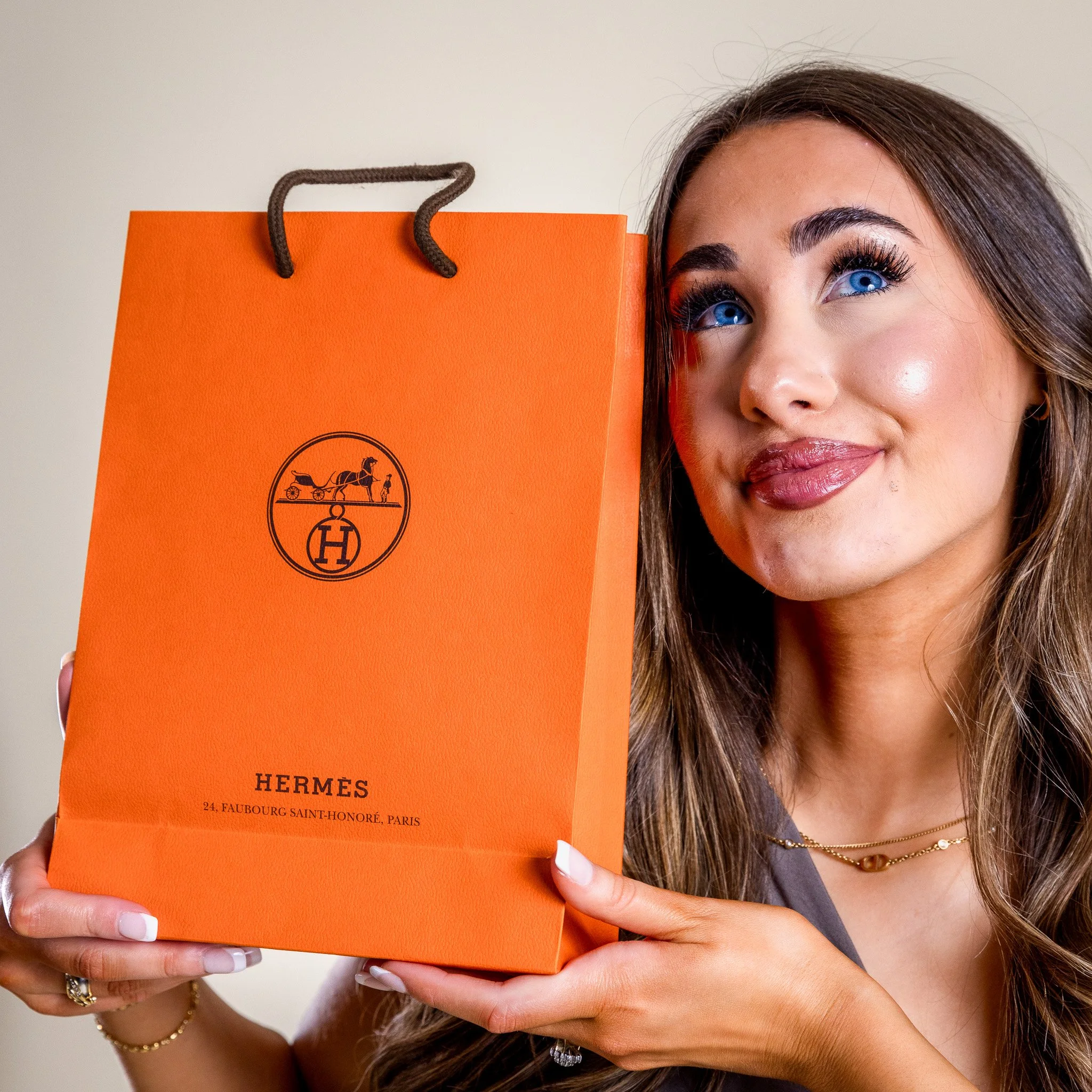 A woman with blue eyes and long brown hair holding an orange Hermès shopping bag with a smile.