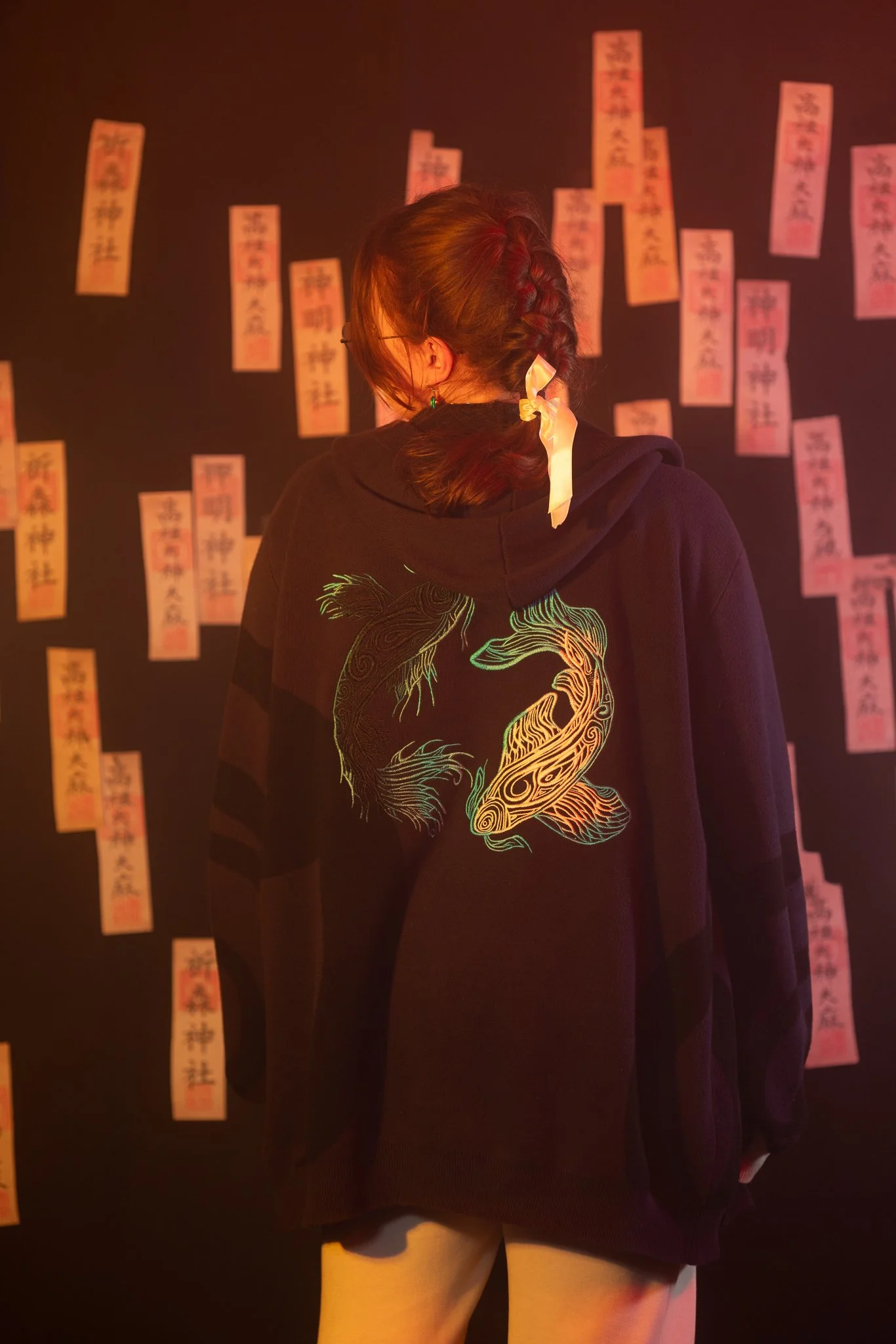 A person with braided hair tied with a white ribbon, wearing a black hoodie with a colorful dragonfish design on the back, observing orange and red paper slips on a black wall in a dim, warm light.