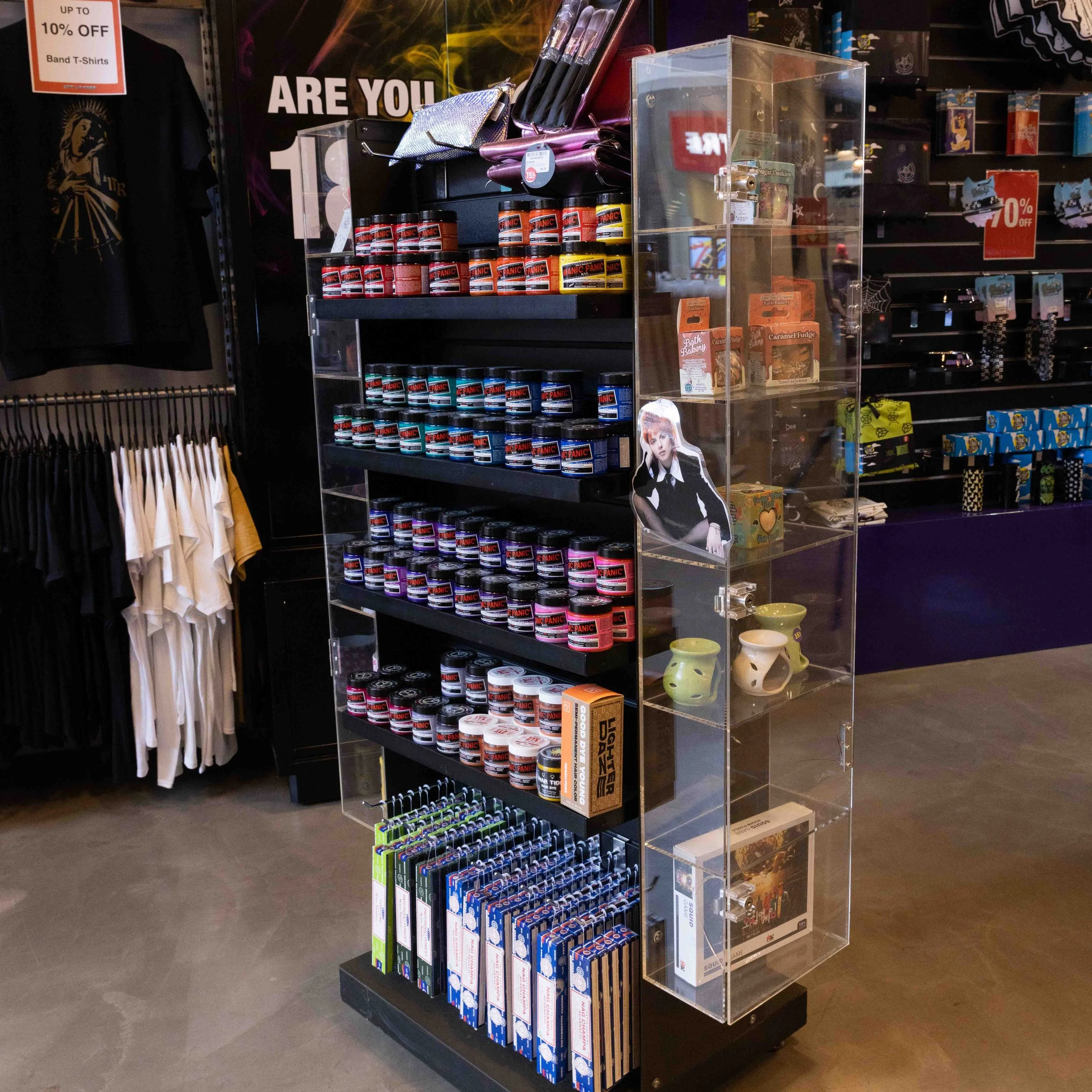A display shelf in a retail store with various small jars of black, red, blue, and pink products, likely cosmetics or small containers, along with some boxed items, figurines, and a cutout photo of a woman. Behind the shelf, there are shirts and hang