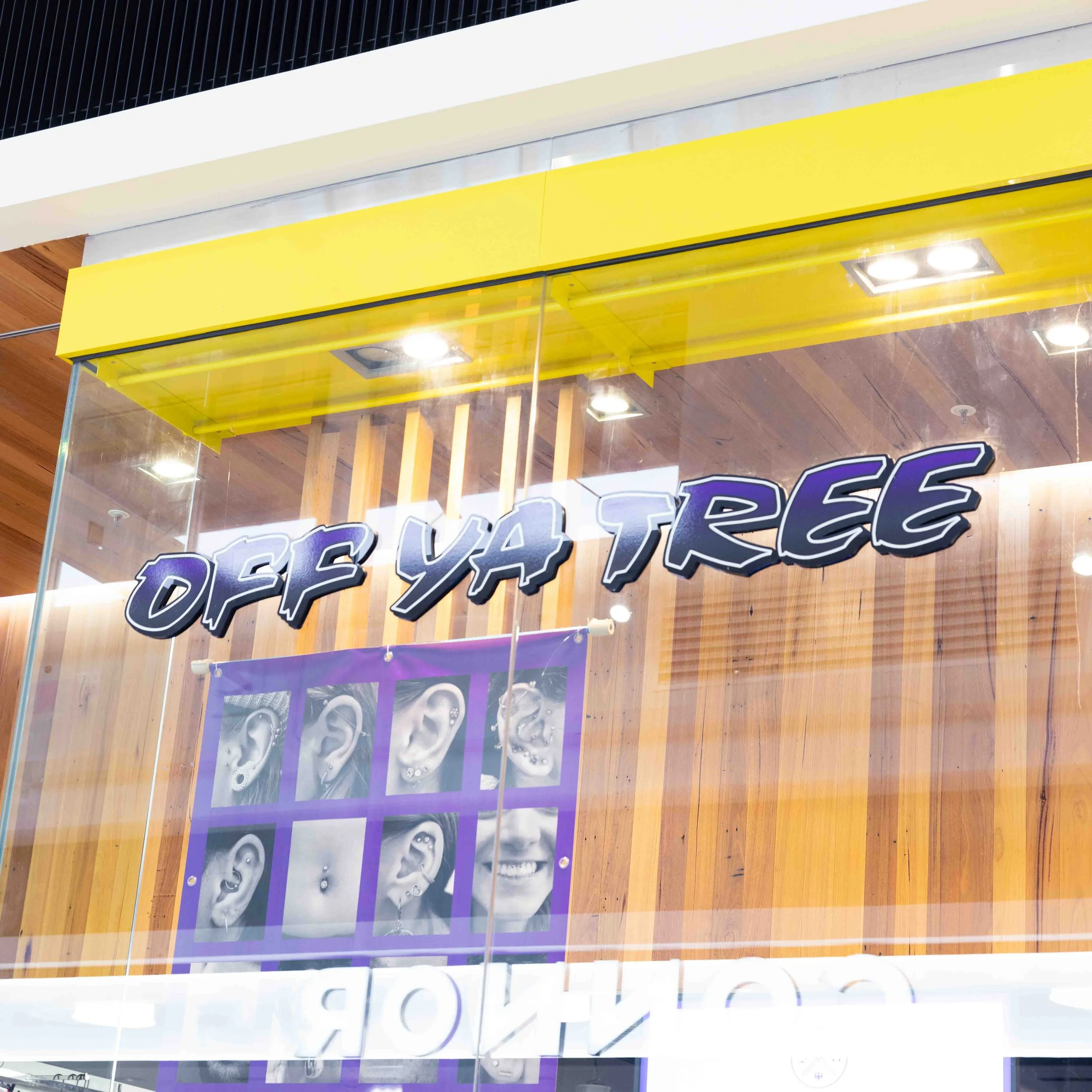 Storefront window with a neon sign that reads 'OFF YA TEEZ', a display of multiple pictures of ears with various piercings, and a yellow awning above.