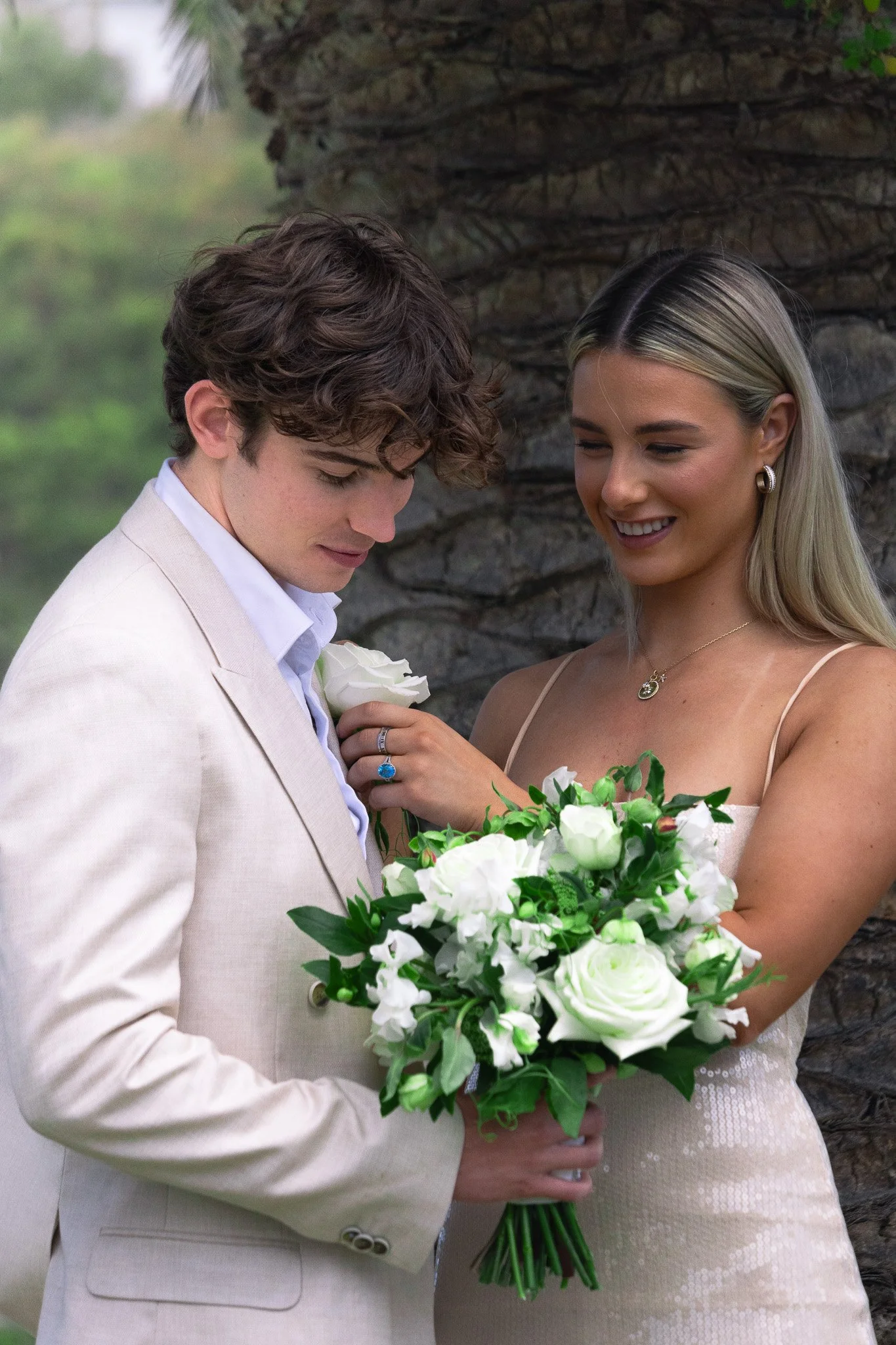 A bride and groom sharing a moment outdoors, the bride holding a bouquet of white flowers and greenery, behind a rocky background.