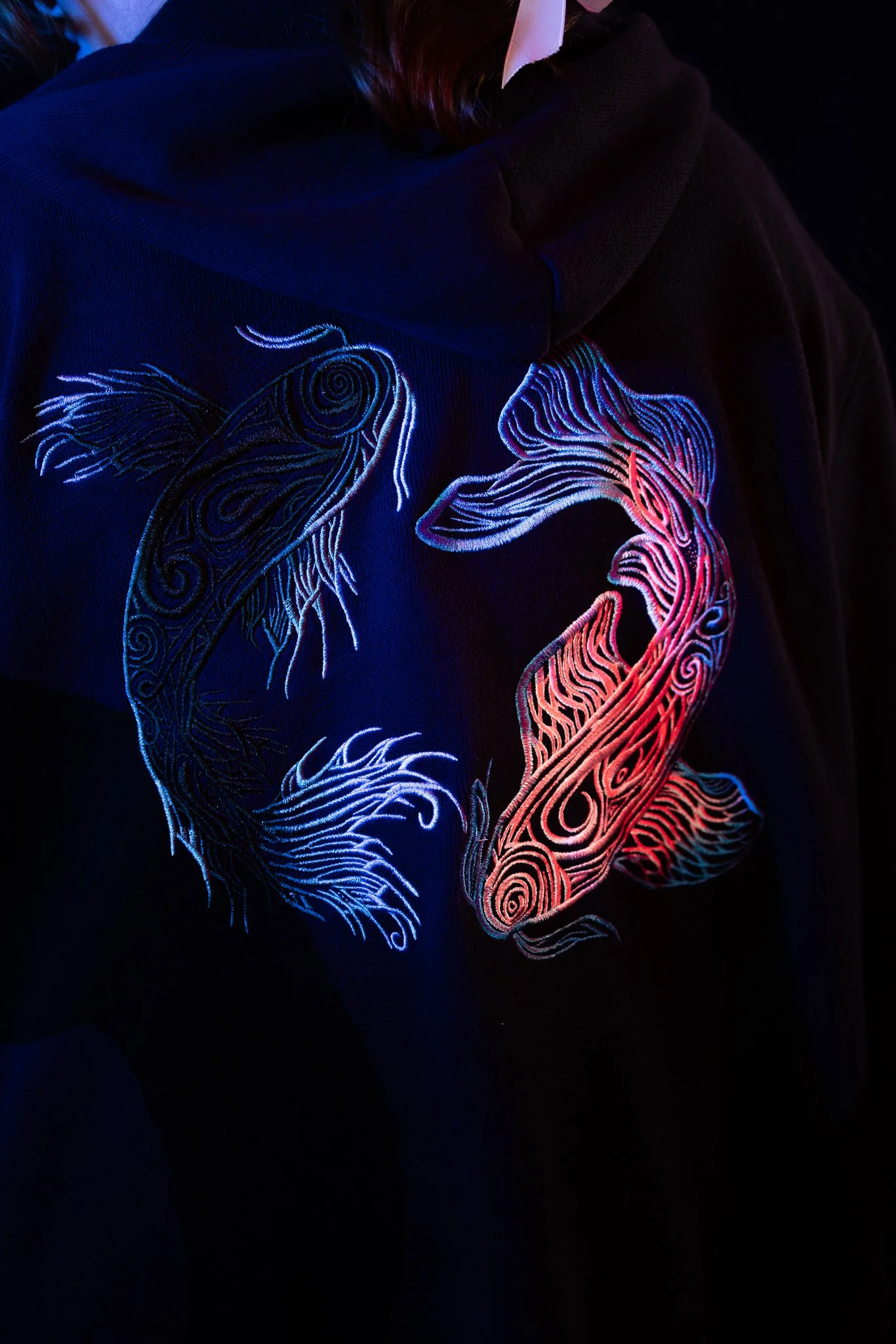 Black hoodie with neon fish embroidery featuring two stylized koi fish, glowing in blue and red, under UV light.