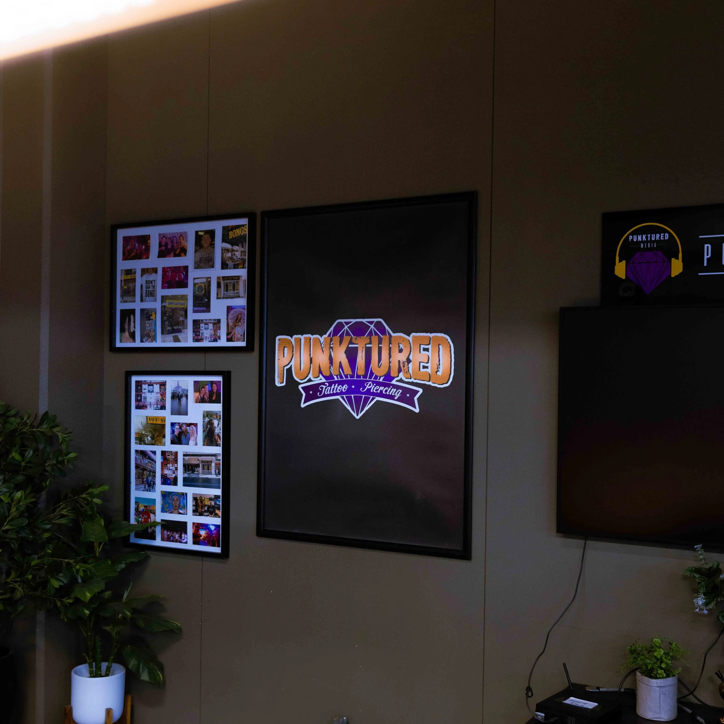 Interior wall with digital signs, including one for 'Punktured' tattoo and piercing shop, with photo collages and potted plants.