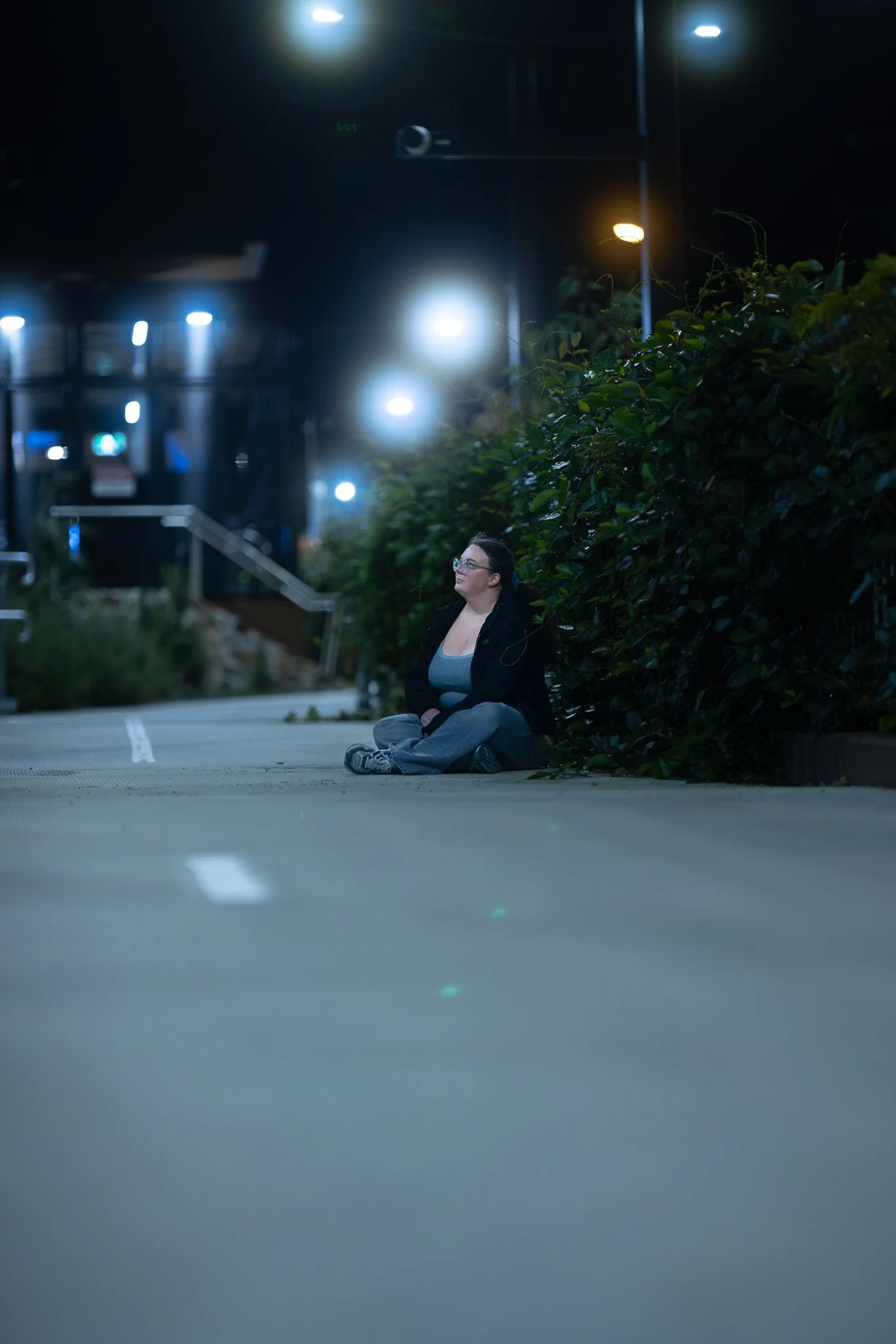A woman sitting alone on a city sidewalk at night, next to bushes, illuminated by streetlights.