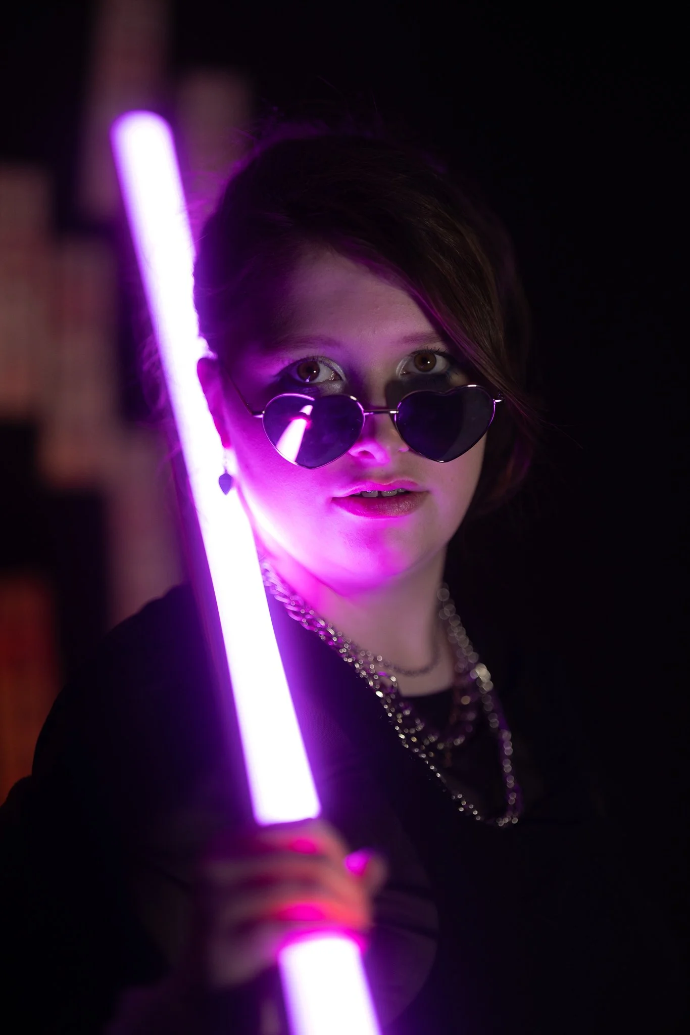 A woman with dark hair, wearing heart-shaped sunglasses and layered necklaces, is holding a purple glow stick in a dark setting, illuminated by neon light.