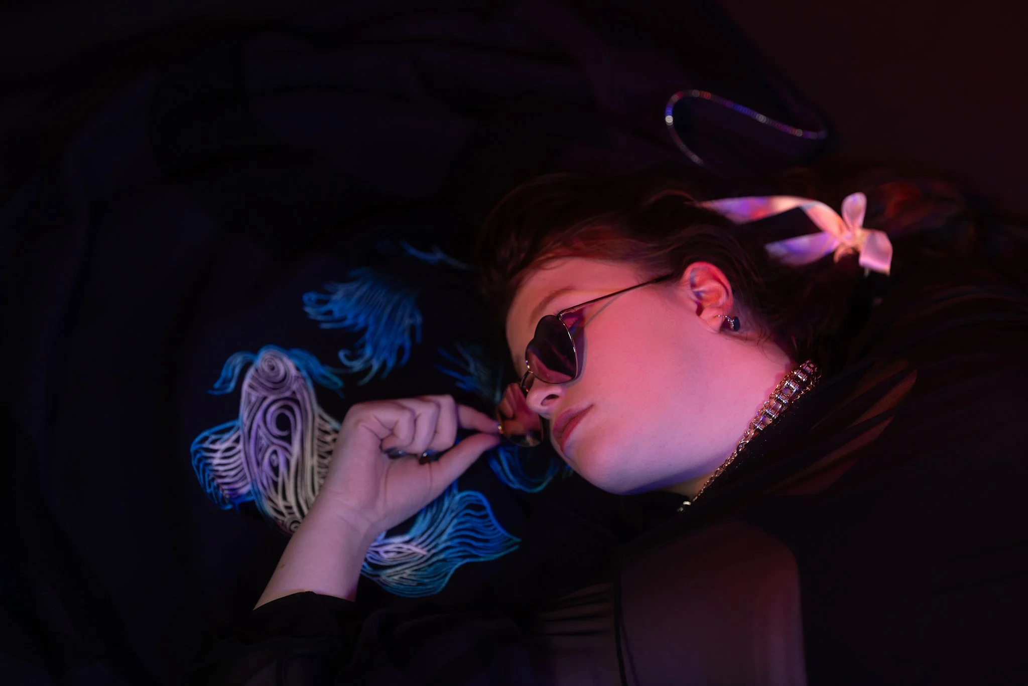 A woman with long dark hair lies on a black surface, wearing black sunglasses, a colorful embroidery on her shirt, and a silver chain necklace. She has a white ribbon in her hair and a black hair tie. The scene is lit with pink and blue lighting.
