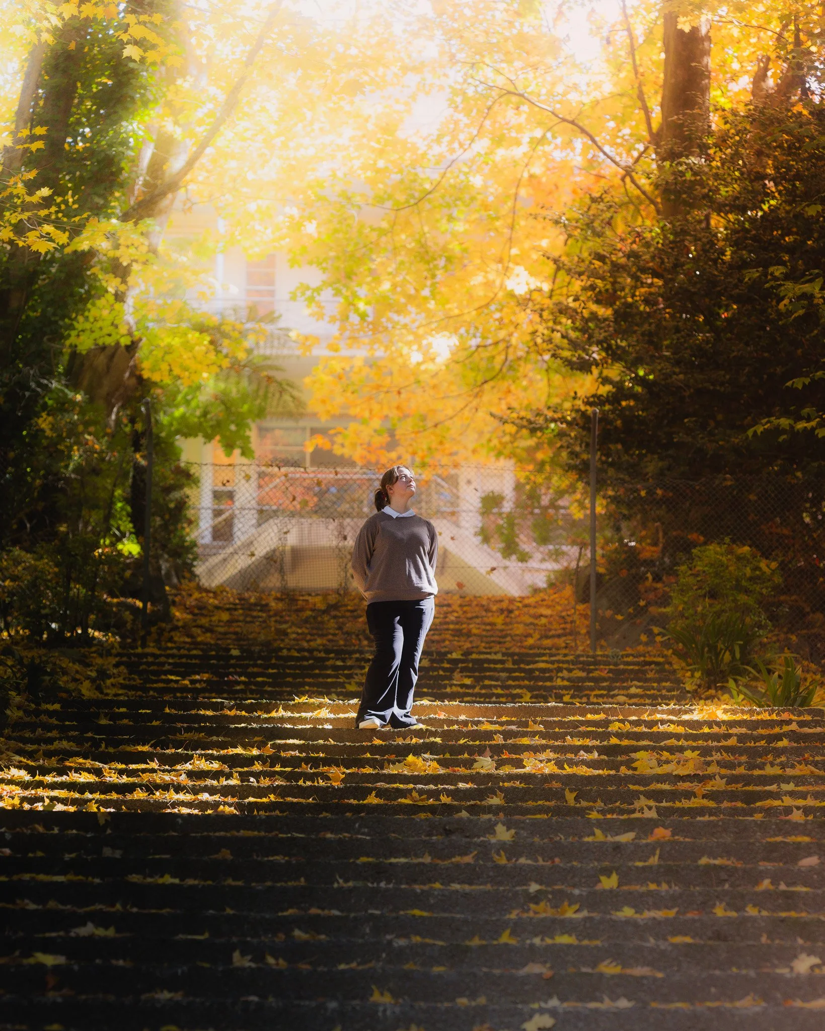 A woman standing on a staircase covered with fallen yellow leaves, surrounded by trees with autumn-colored foliage, illuminated by warm sunlight in a park or garden.