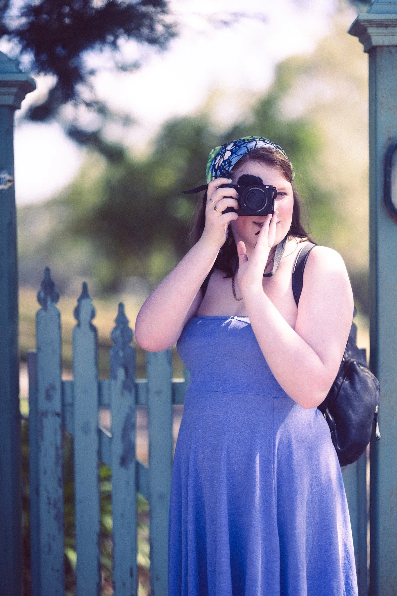 A woman wearing a blue dress and a floral headscarf, taking a photo with a camera, standing in front of a blue picket fence.
