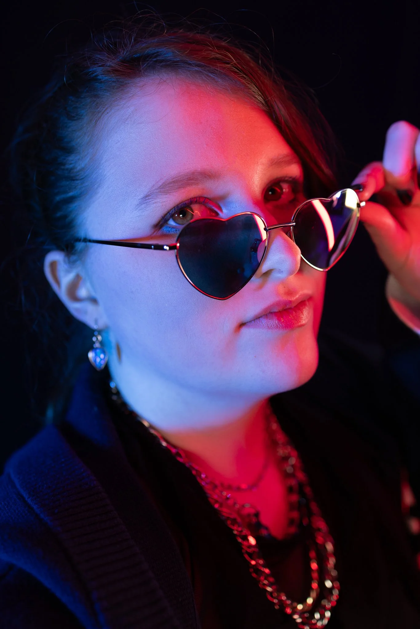 A young woman with brown hair, wearing heart-shaped sunglasses, jewelry, and dark clothing. She is holding her sunglasses with her right hand and looking at the camera, with a colorful lighting effect.