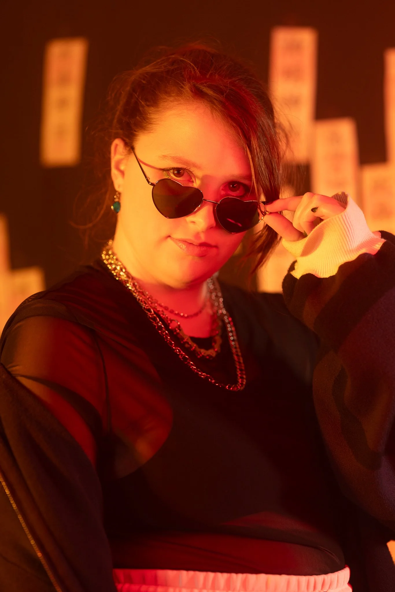 A young woman wearing sunglasses, earrings, and layered necklaces, with fashionable clothing, looking at the camera with a confident expression, posed against a background with blurred text.