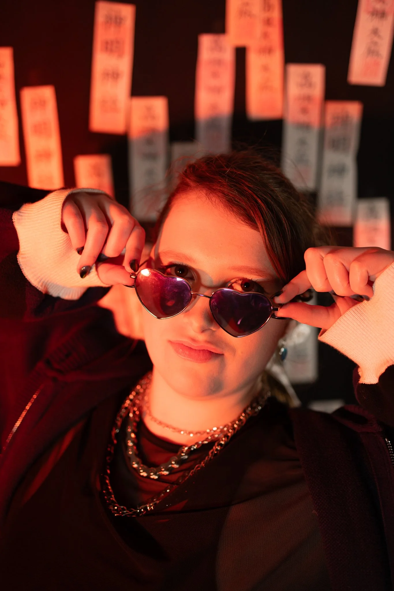 A young woman with dark hair and rings on her fingers is wearing heart-shaped sunglasses with pink lenses, holding them with both hands, and looking directly at the camera. She is dressed in dark clothing with layered necklaces, and there are hanging