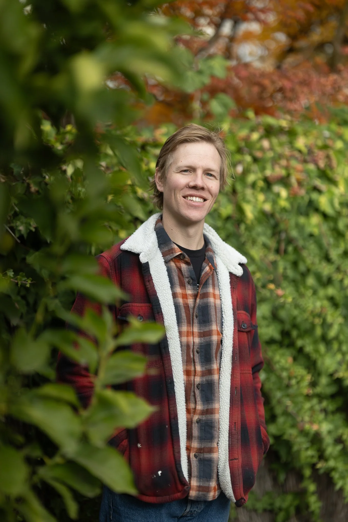 A young man with blonde hair smiling outdoors among green and red autumn foliage, wearing a plaid shirt and a red jacket with a fleece lining.