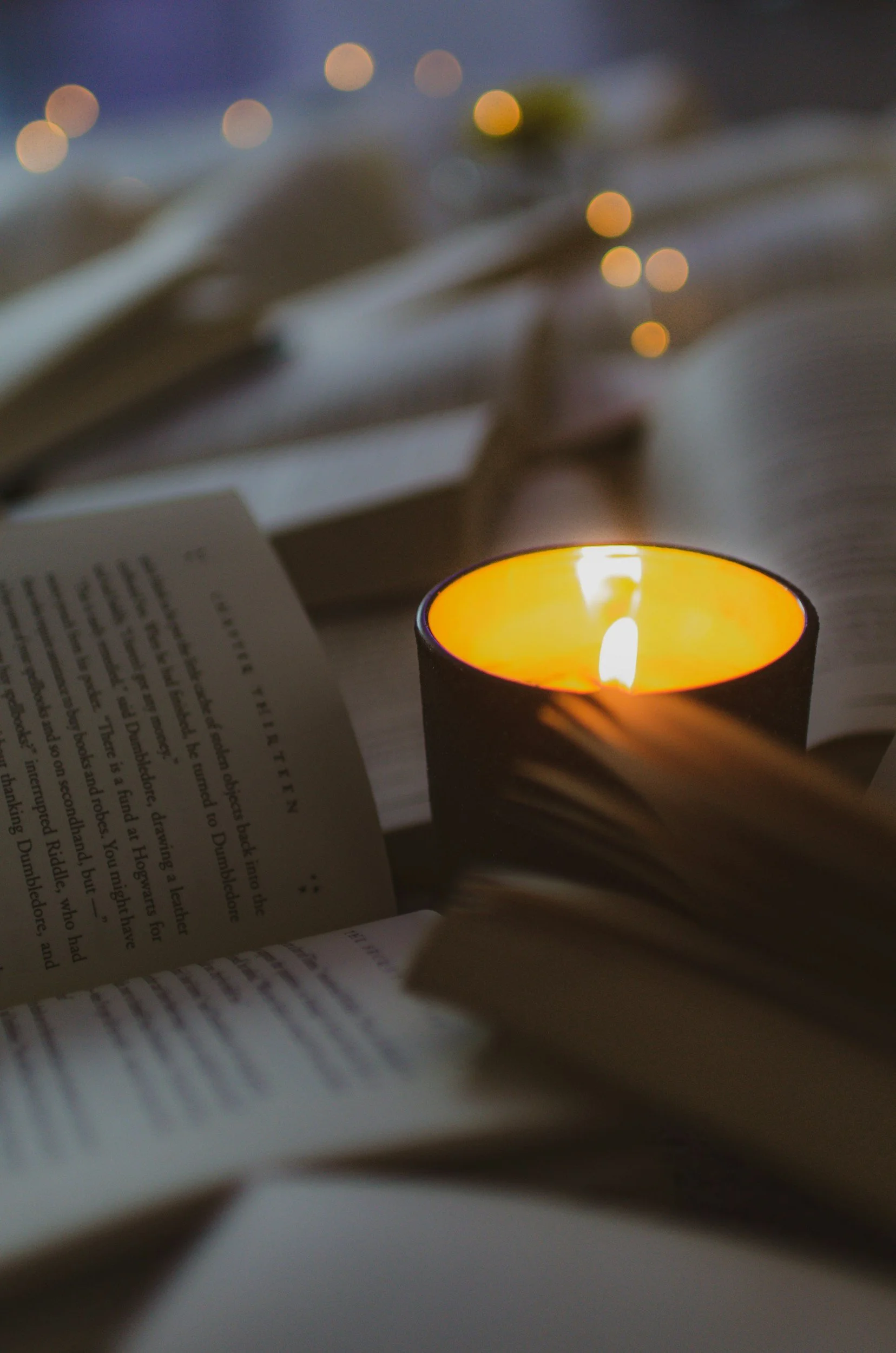 Blurred open book with text in the foreground, a lit candle with an orange glow, and blurred string lights in the background creating a cozy atmosphere.