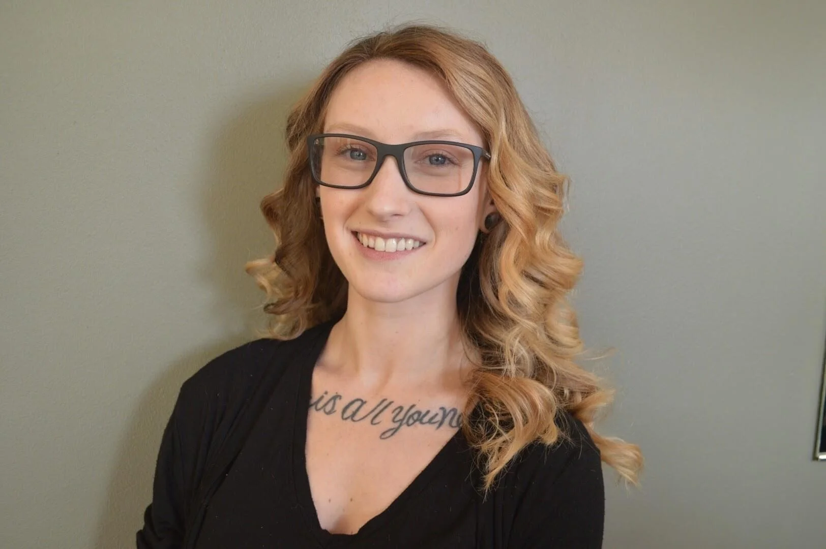 Close-up of a young woman with curly blonde hair, wearing black glasses and a black top, smiling against a plain wall background.