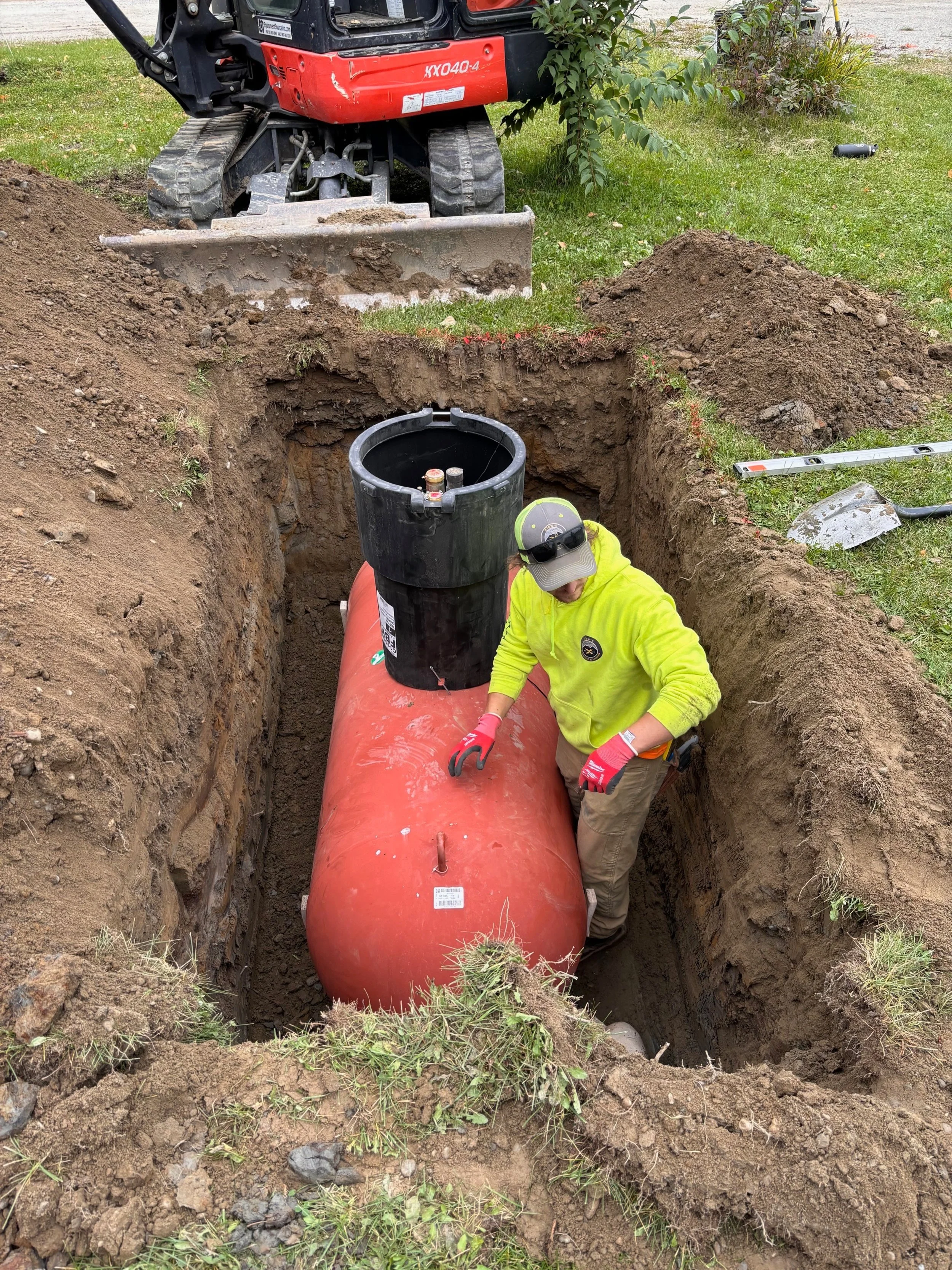 A worker installs a large orange underground tank in a deep trench, with a mini excavator nearby on a grassy lawn.