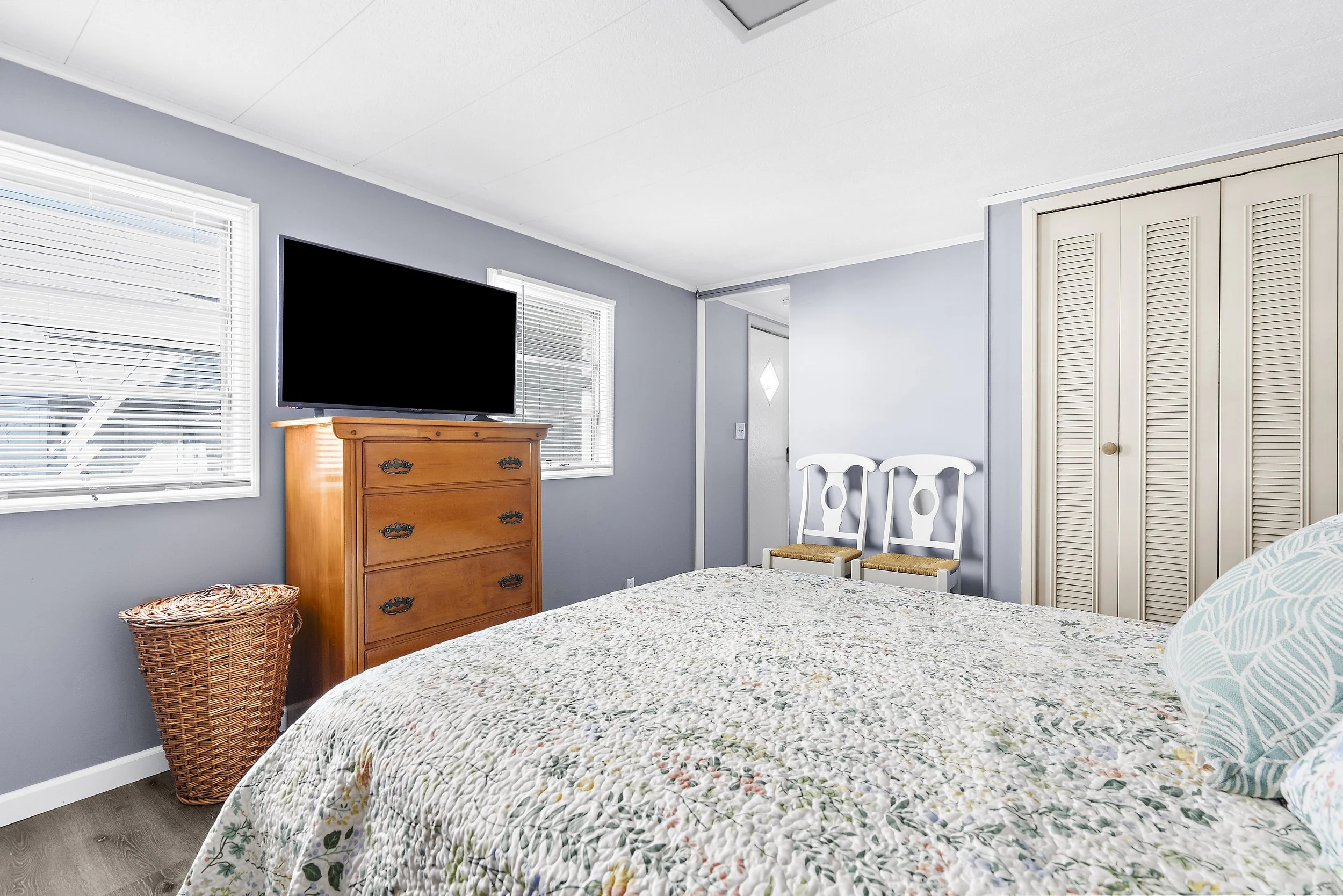 A bedroom with a bed, two white chairs, a wooden dresser with a black TV, window blinds, and a closet with louvered doors.