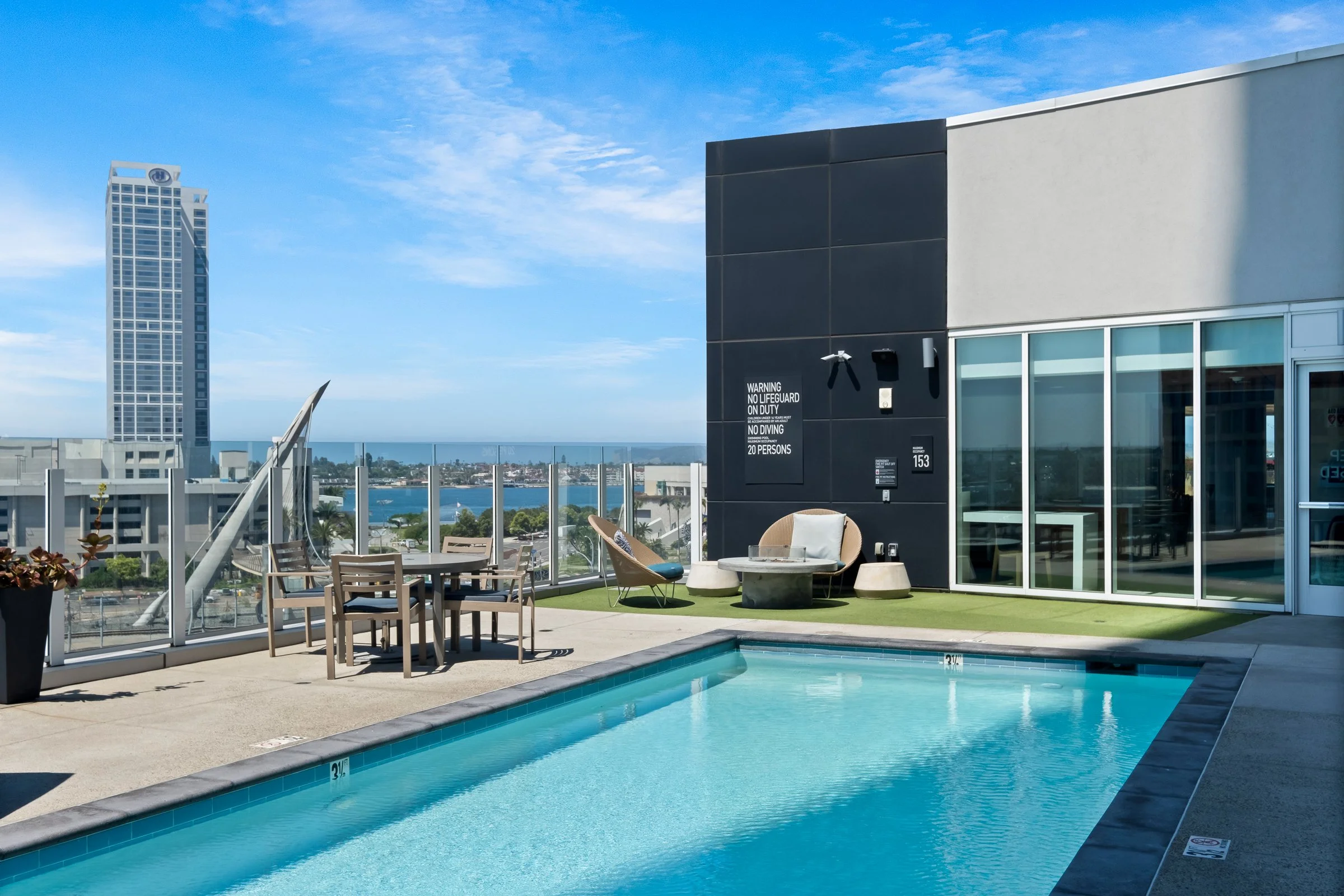 Rooftop swimming pool with outdoor furniture, city skyline, and ocean view in the background.