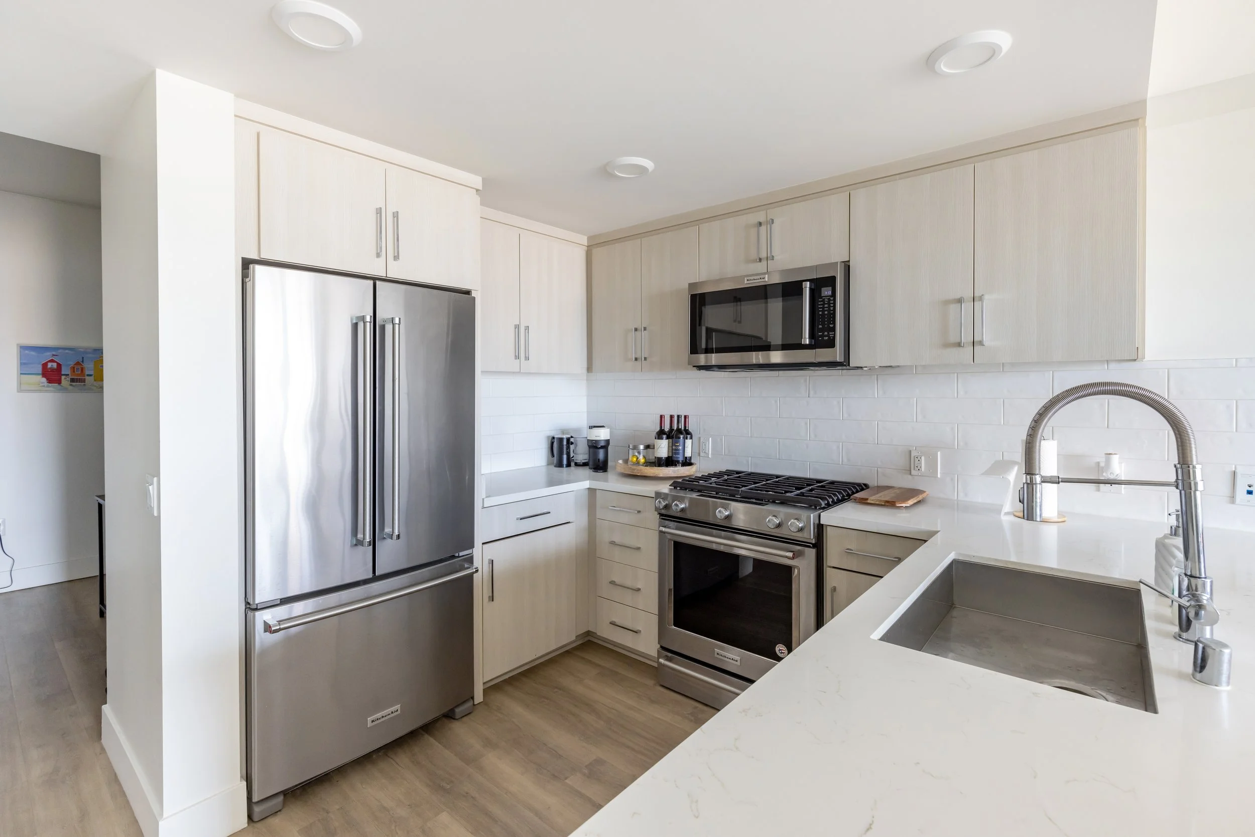 Modern kitchen with stainless steel refrigerator, microwave, oven, and sink, light wood cabinets, white countertops, and a white subway tile backsplash.