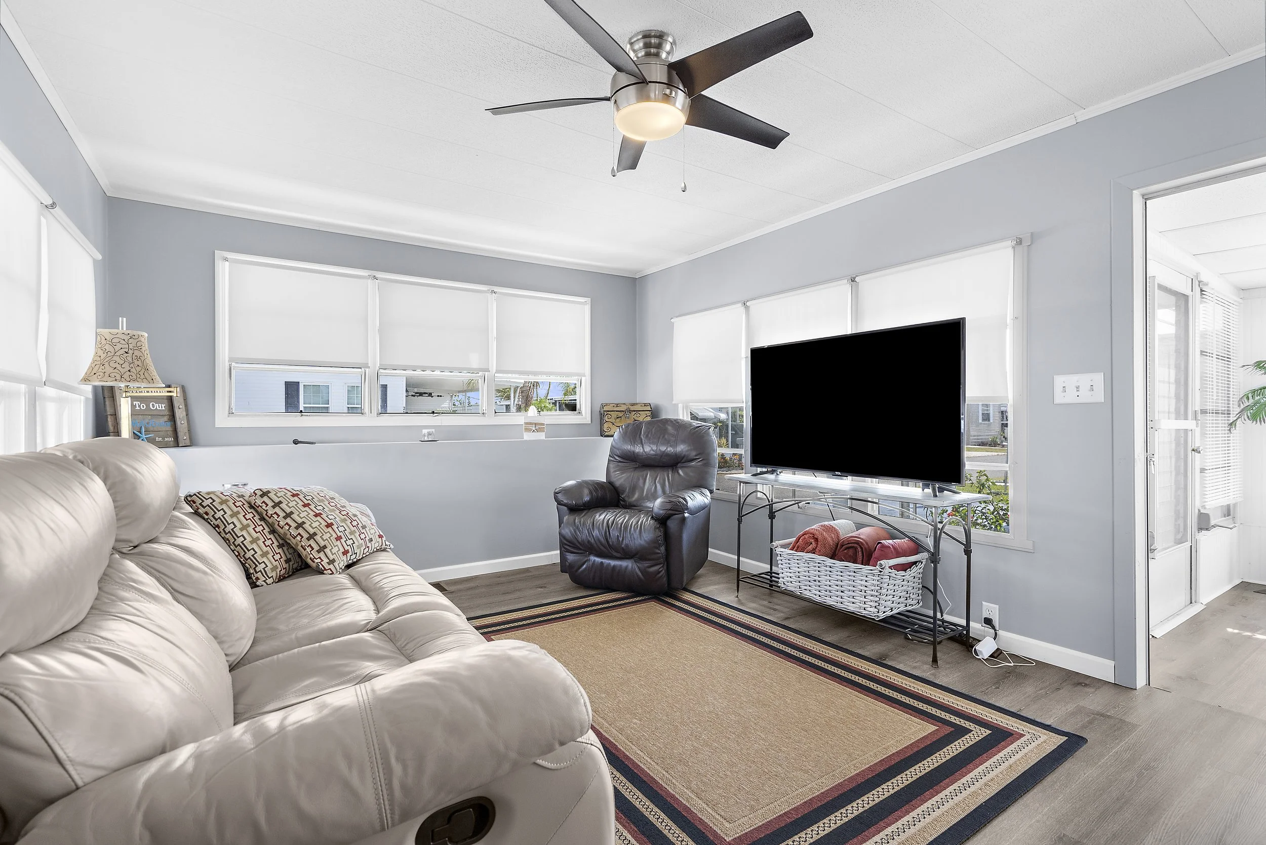 Living room with a cream leather sofa, a dark leather armchair, a TV on a stand with folded towels underneath, a beige area rug, and windows with white blinds, in a home with light blue walls.