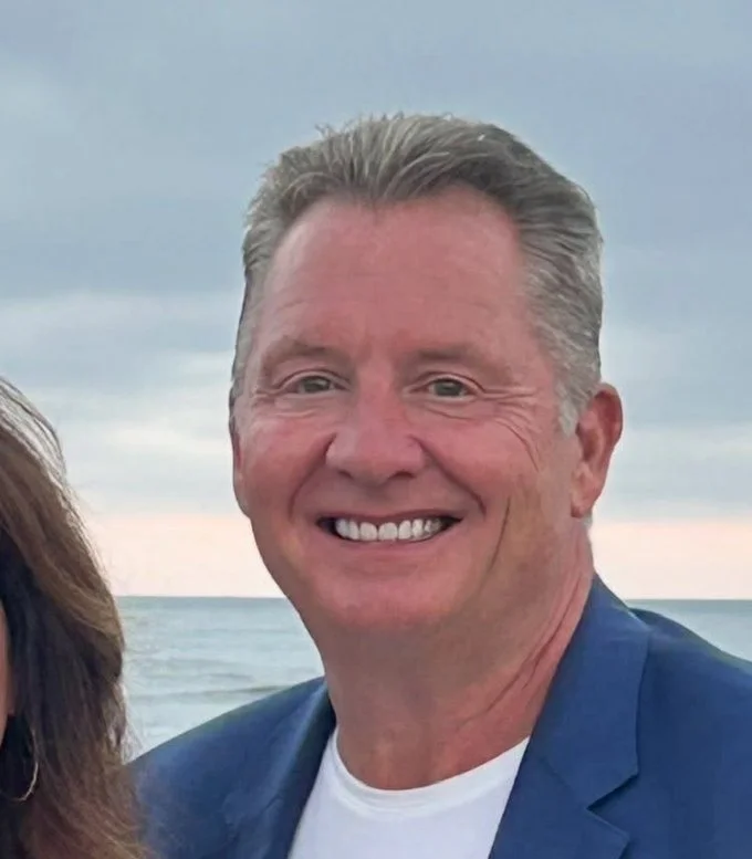 Smiling man with light-colored hair, wearing a blue blazer and white shirt, standing outdoors near water with cloudy sky in background.