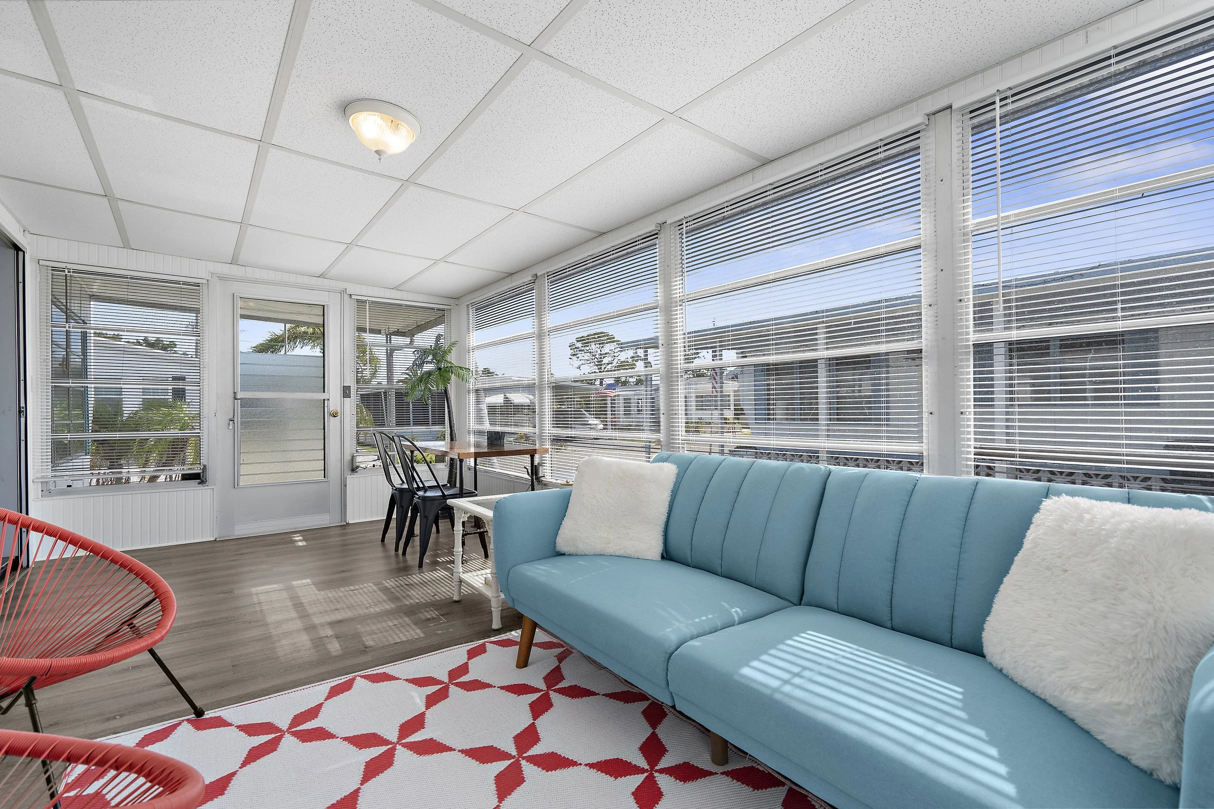 Bright sunroom with large windows and blinds, blue sofa with white pillows, red and white geometric rug, black metal chairs, small wooden table, potted plant, view of neighboring houses and trees outside.