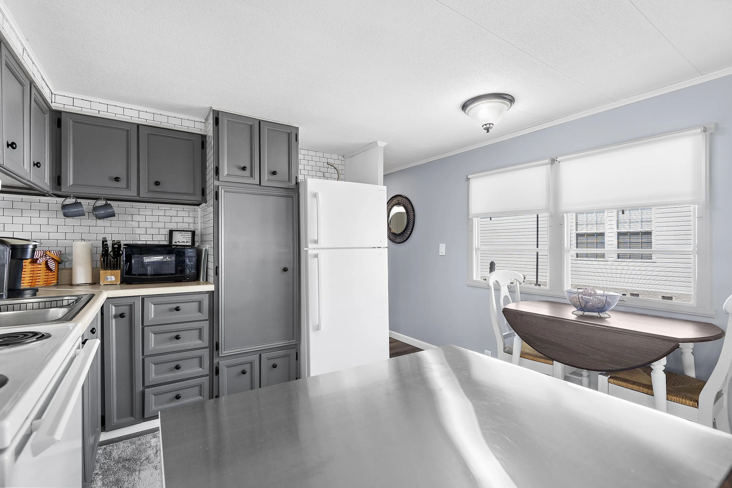 Kitchen with gray cabinets, white subway tile backsplash, white refrigerator, and a small dining area with a table and two chairs near windows.