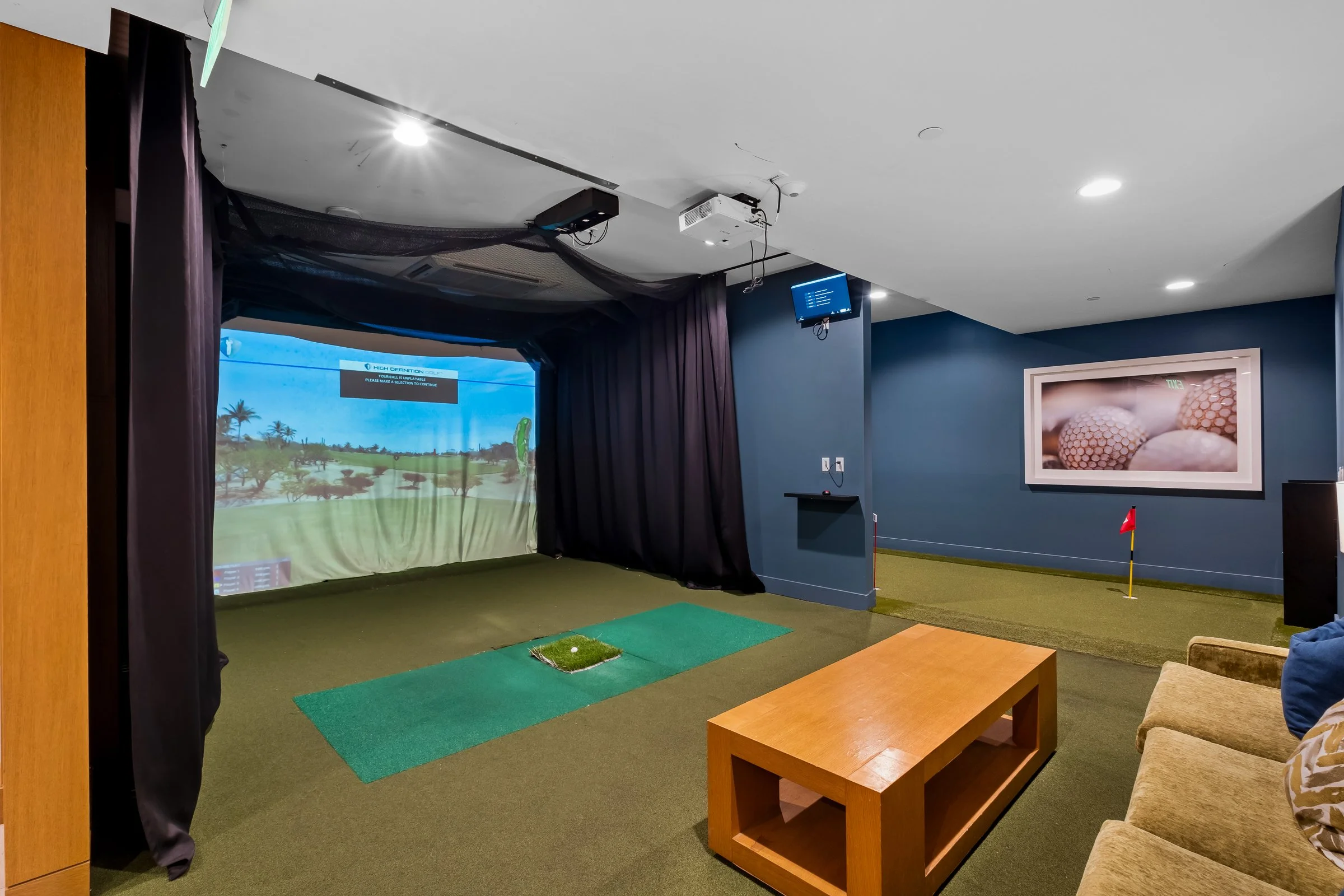 Indoor golf simulation room with a large screen displaying a golf course, green turf mat with a golf ball, beige sofas, a wooden table, and golf-themed wall art.