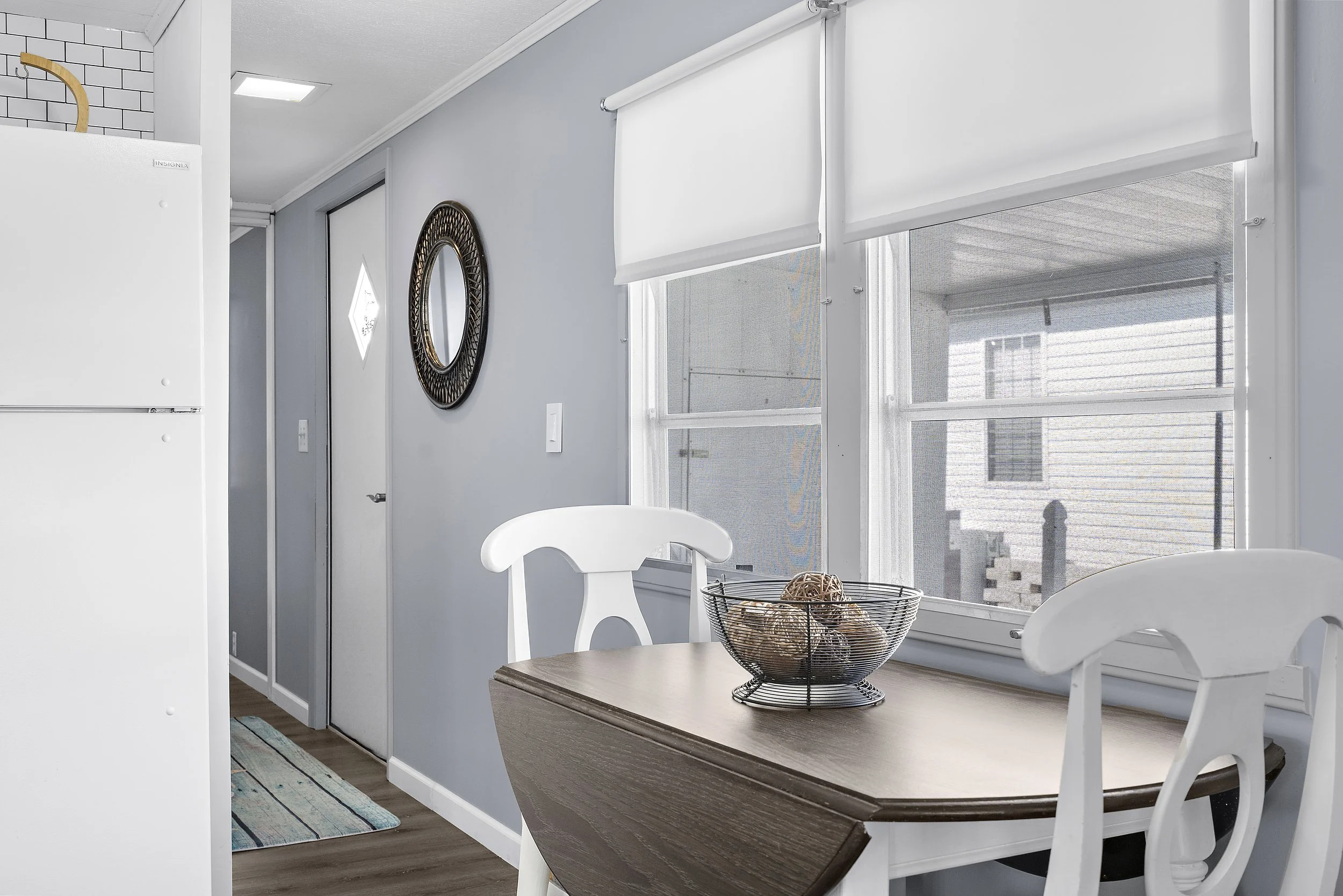 A dining area near a window with white blinds, a wooden table with white chairs, a wire bowl with decorative orbs, and a wall mirror.