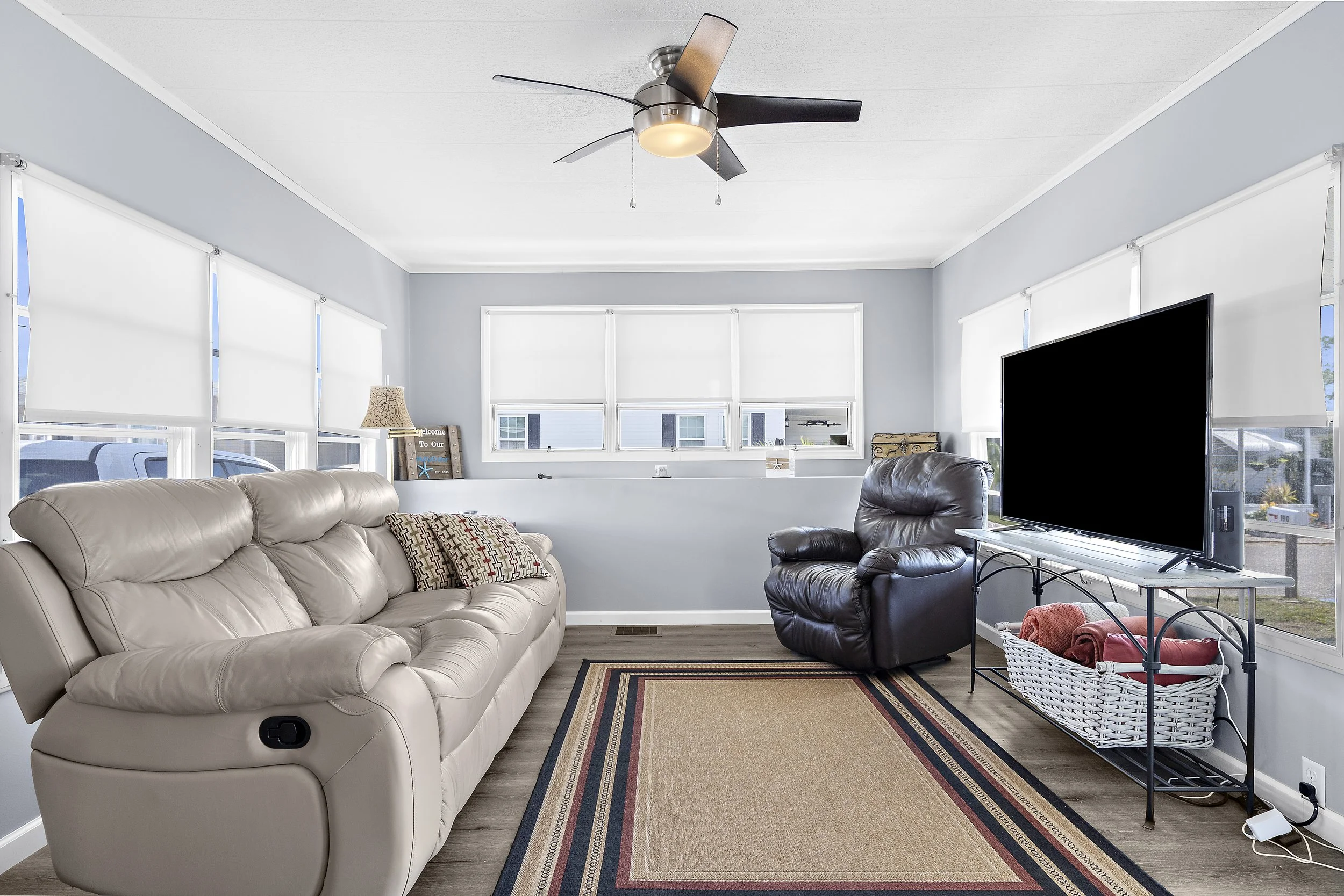 Living room with light blue walls, beige leather sofa, black leather recliner, large flat-screen TV, white blinds on windows, a small table with a basket of blankets, a decorative sign, and a ceiling fan