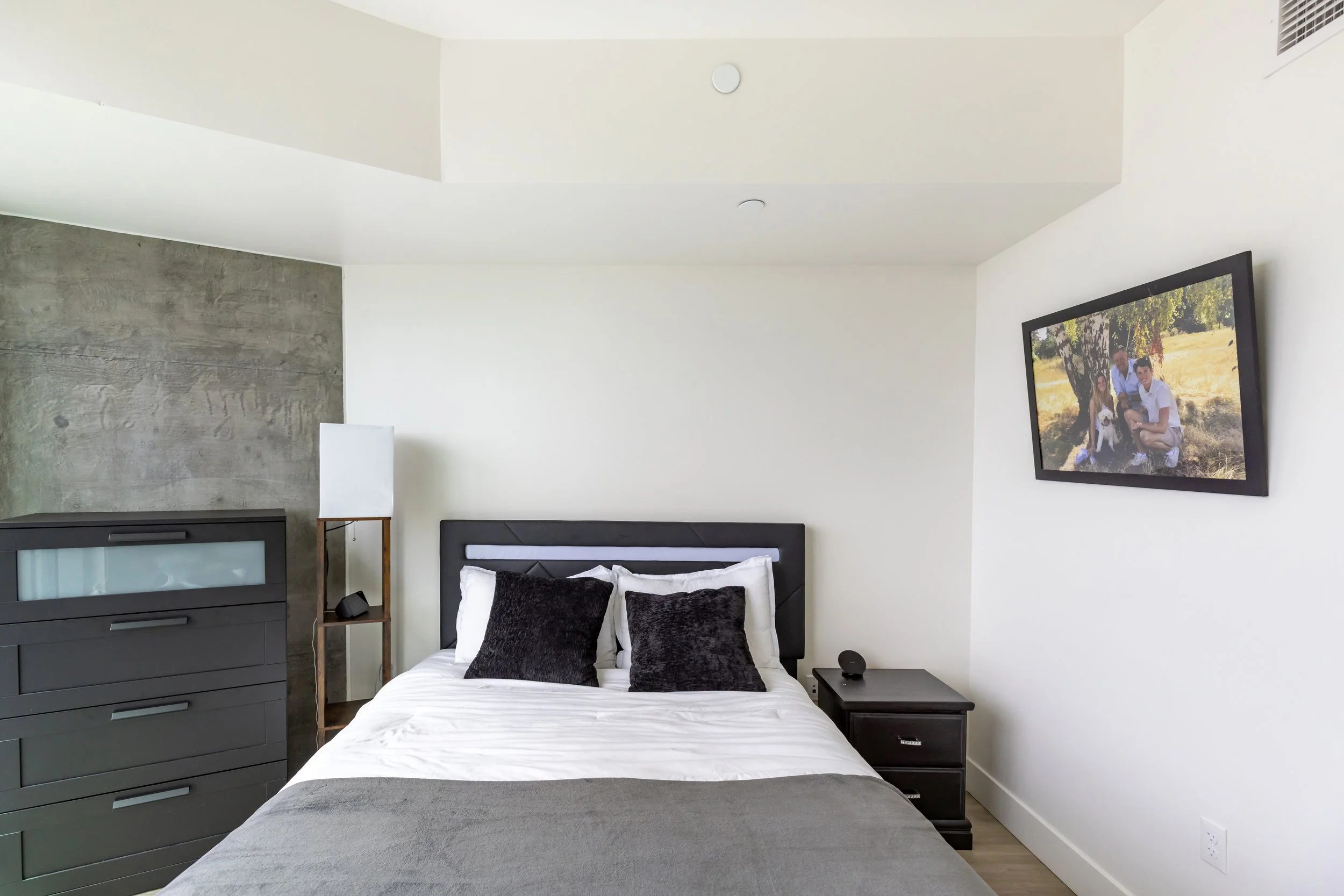 Modern bedroom with a bed, black pillows, nightstand, dresser, a tall lamp, and a wall-mounted TV displaying a family photo, with white walls and a textured accent wall.