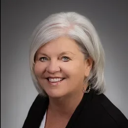 A smiling middle-aged woman with short white hair, wearing a black blazer and earrings, against a gray gradient background.