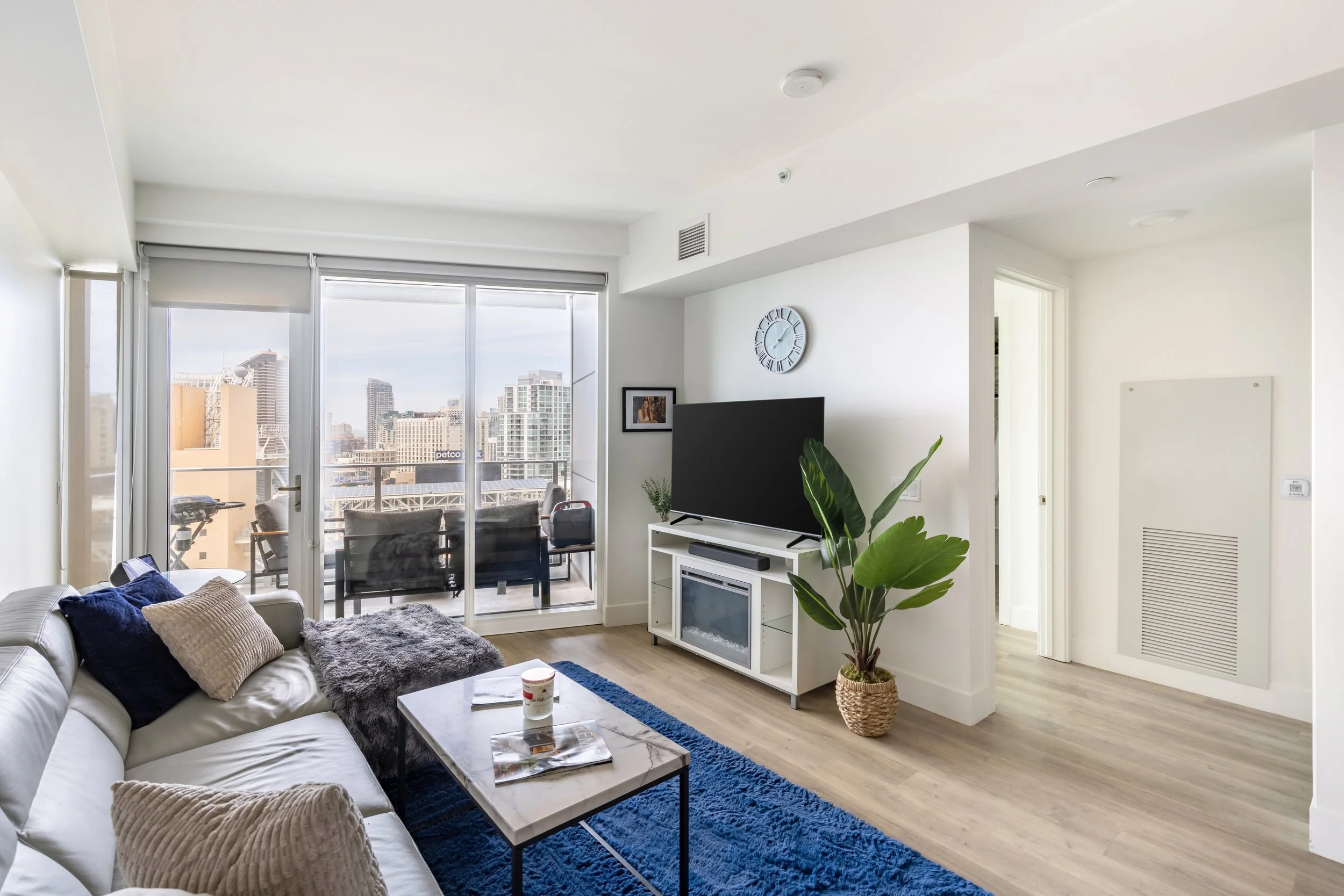 Modern living room with a white sofa, blue rug, and coffee table. Large glass sliding door opens to a balcony with city skyline view. Flat-screen TV on white stand, potted plant nearby, and a wall clock above the TV. Bright, minimal decor with white