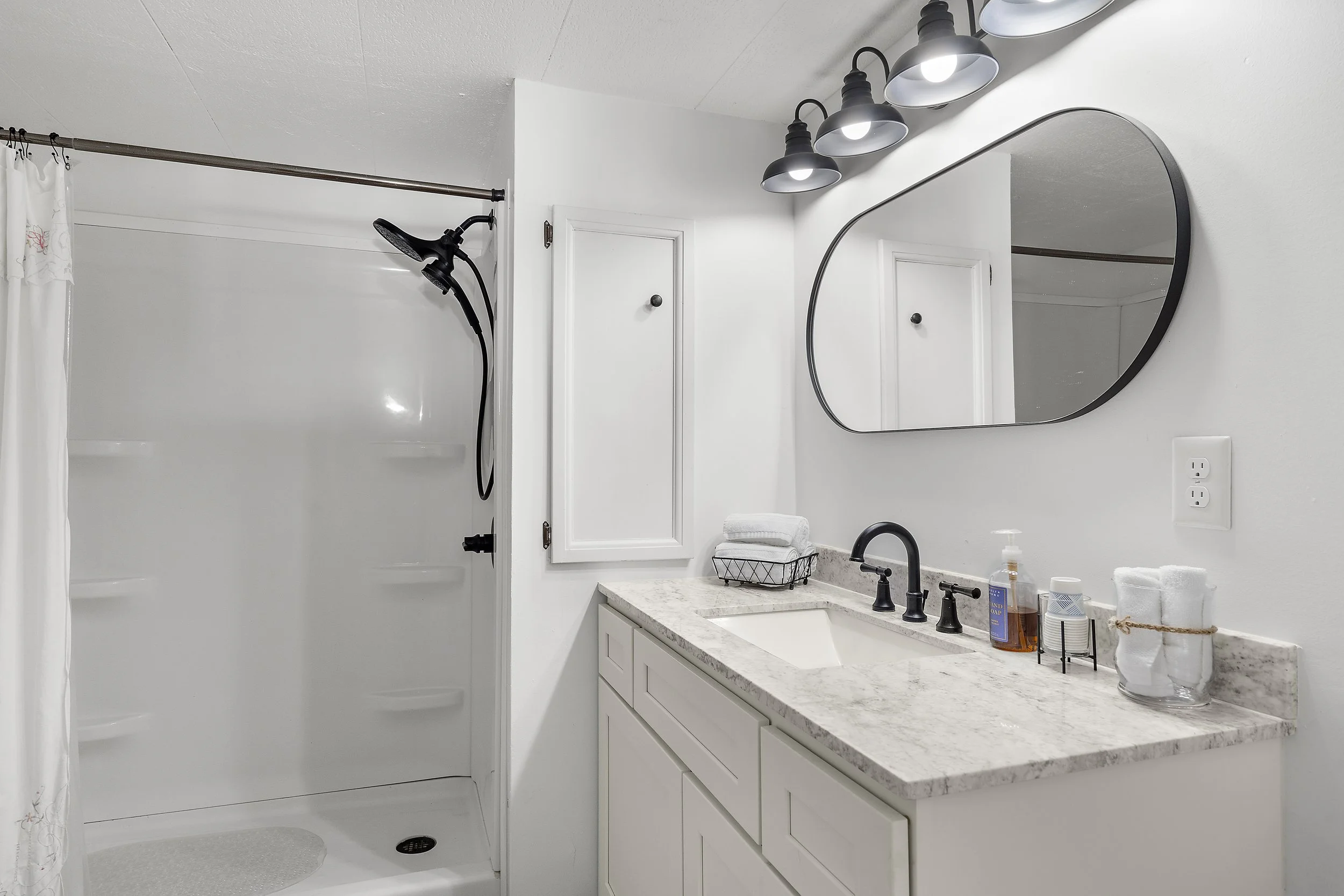 Bathroom with walk-in shower on the left, white walls, a countertop with a sink, black faucet, and various toiletries, a modern mirror above the sink, and a row of four black-shaded wall lights.