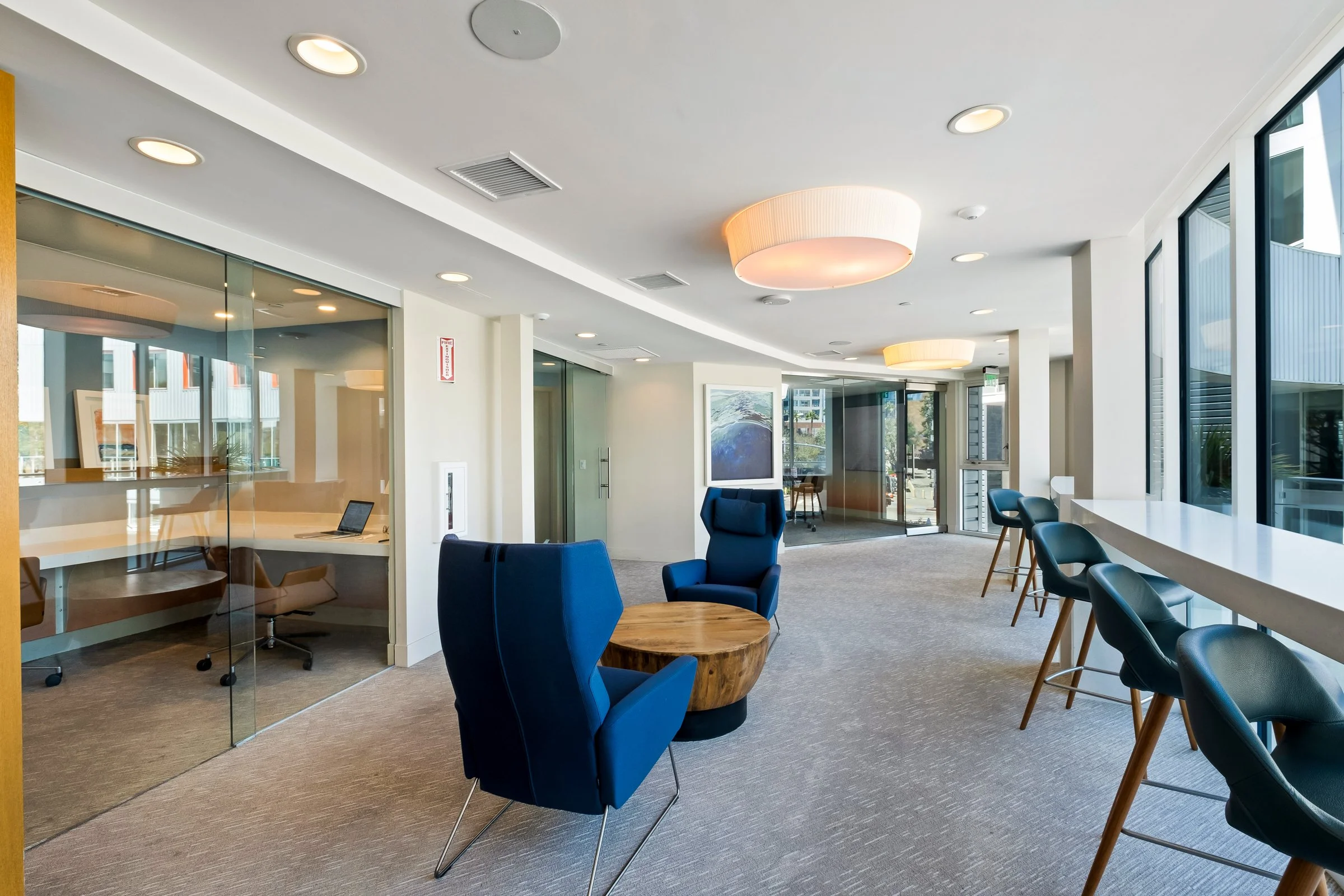 Modern office lounge area with blue armchairs, a round wooden table, bar-style seating by large windows, and a meeting room with glass walls.