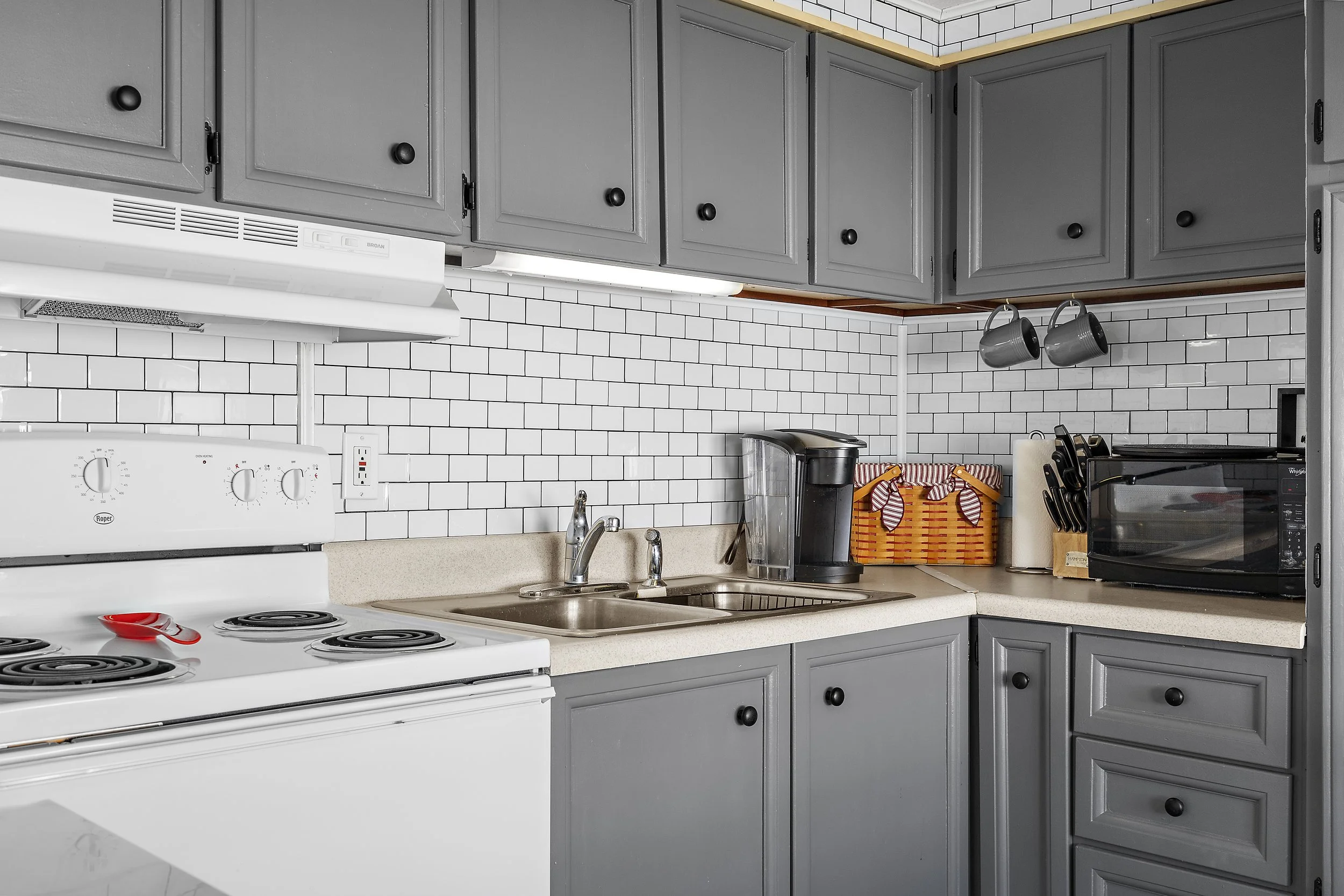 Kitchen with gray cabinets, white subway tile backsplash, beige countertop, stove, double sink, coffee maker, toaster oven, hanging mugs, paper towel roll, and orange baskets.