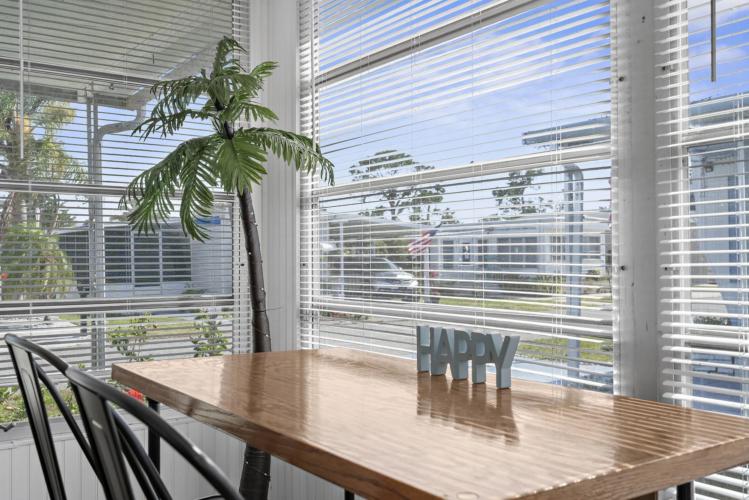 Sunlit dining area with wooden table, metal chairs, large potted palm tree, and decorative letters spelling 'HAPPY' on the table. Large window with blinds and street view outside.