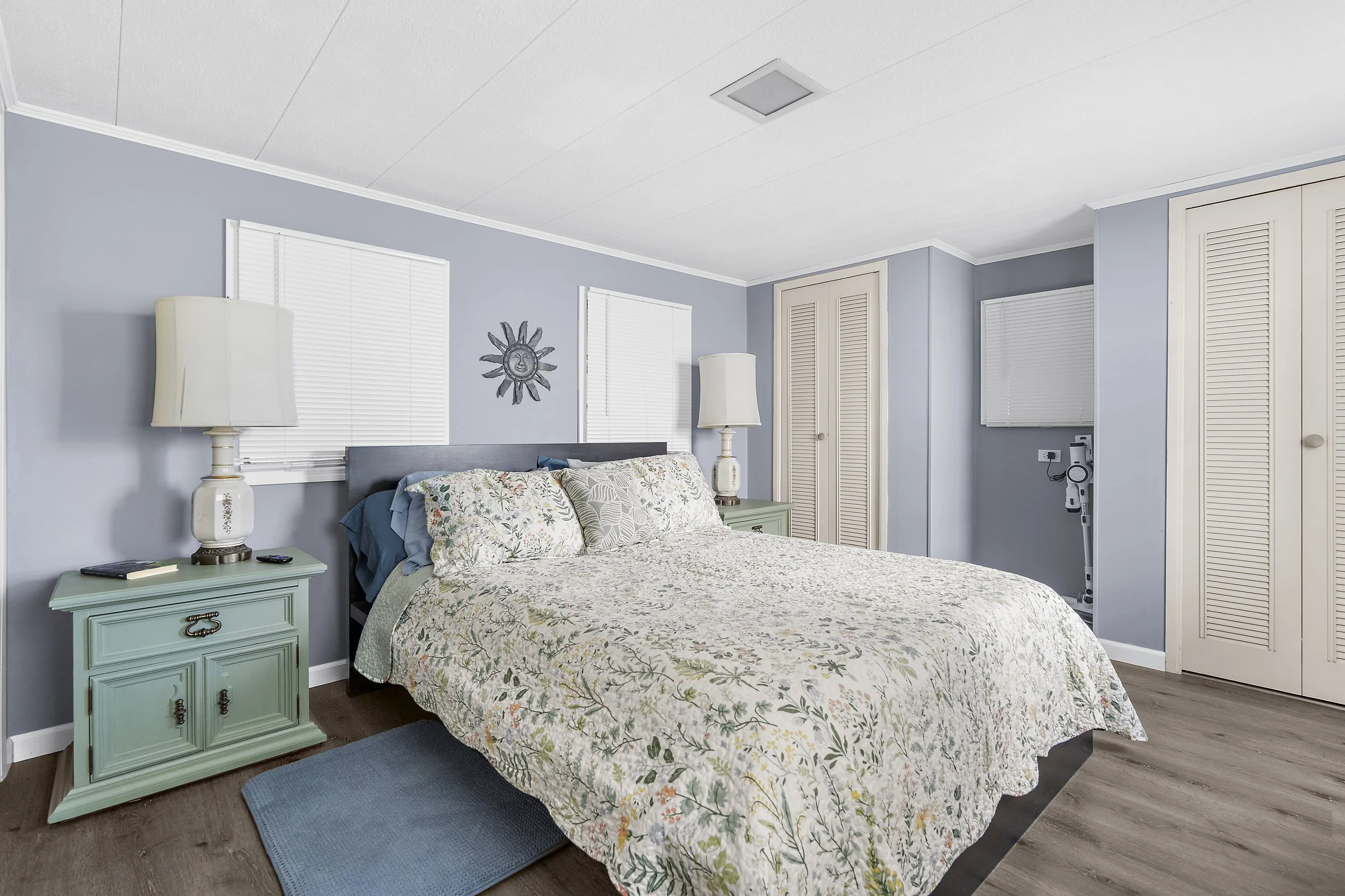 A cozy bedroom with a light blue wall, a bed with floral bedding, two nightstands with white lamps, and a closet with louvered doors.