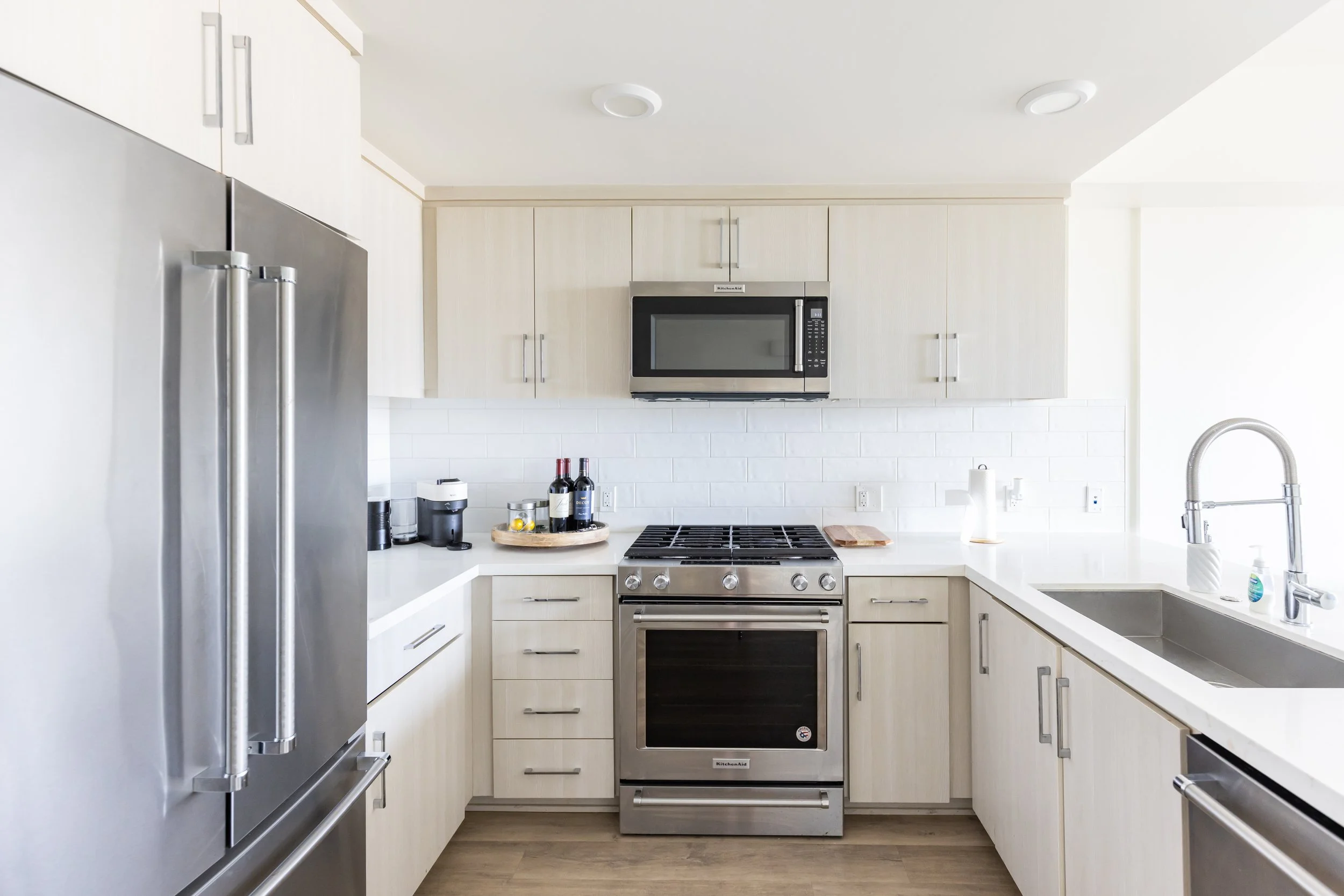 Modern kitchen with stainless steel refrigerator, oven, microwave, and sink. White cabinetry, wood floors, and white subway tile backsplash.