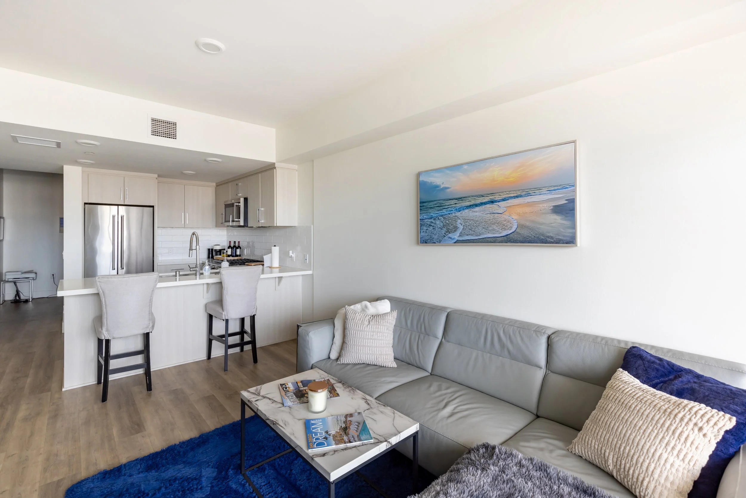 Living room with beige leather sofa, blue rug, and a marble coffee table, adjacent to an open kitchen with white cabinets, stainless steel refrigerator, and a wall-mounted TV showing a beach scene