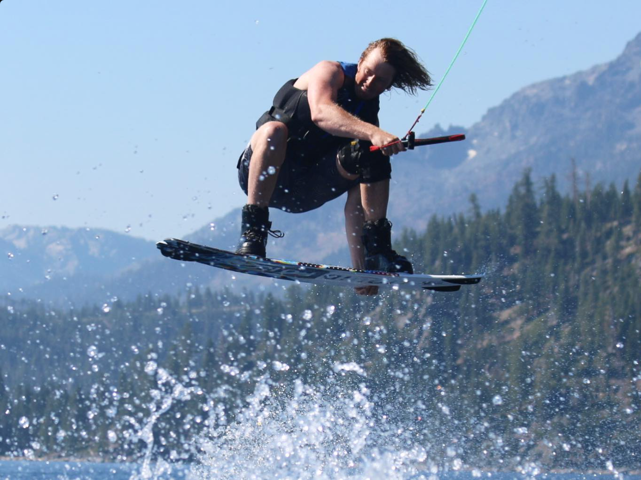 Person wakeboarding on water with mountains in the background, wearing a life vest and holding a handle.