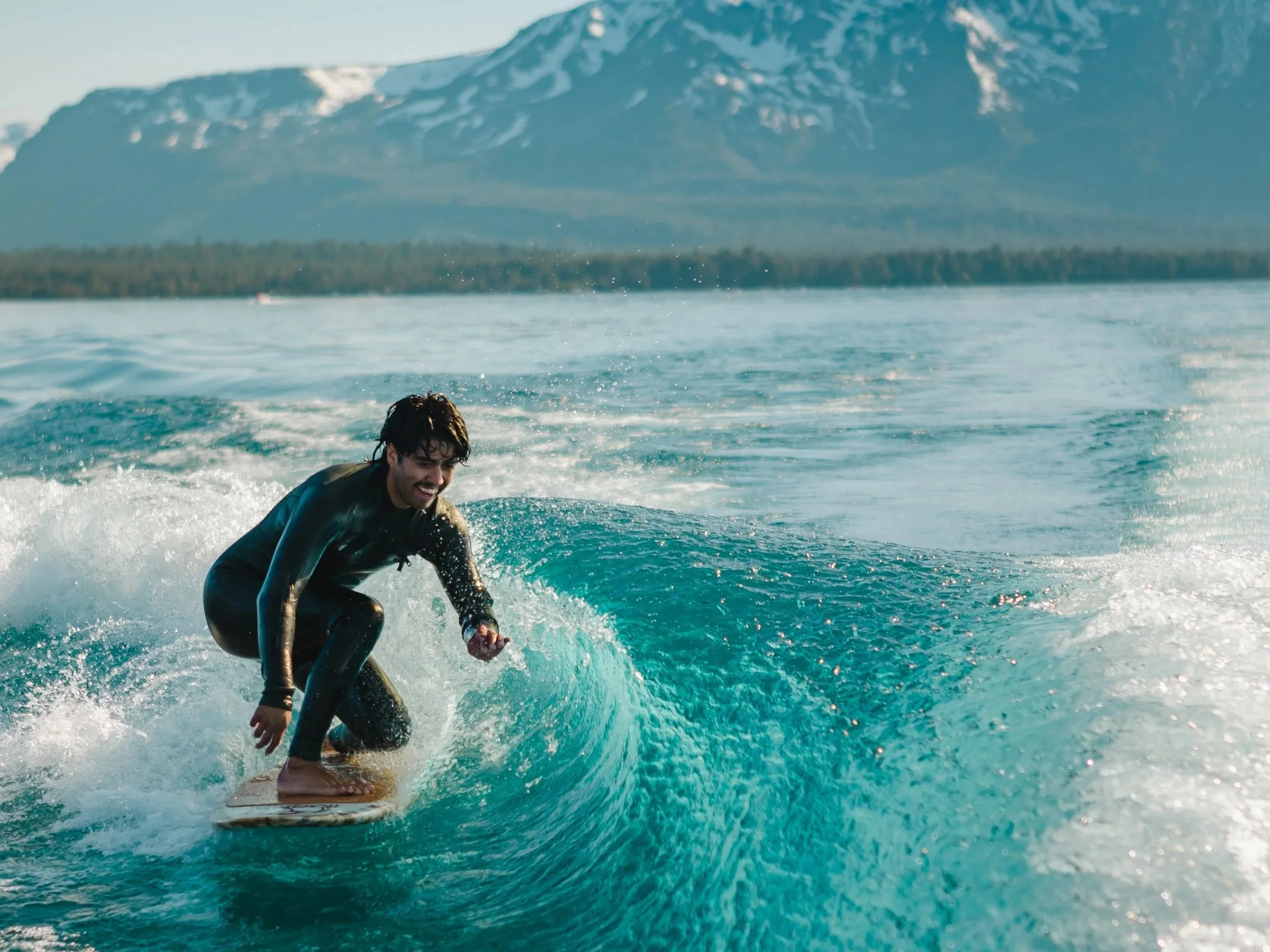 A young man surfing on a wave near the shore with snow-capped mountains in the background.